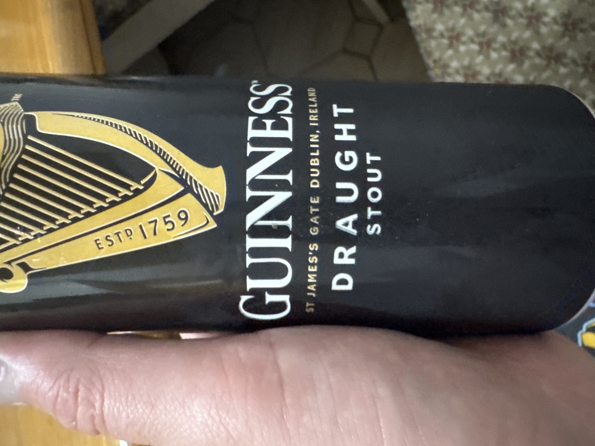 A person's hand is holding a black can of Guinness Draught Stout. The can has the Guinness harp logo in gold and the text "GUINNESS," "ST JAMES'S GATE DUBLIN, IRELAND," and "DRAUGHT STOUT" printed in white. The background is blurred and shows a patterned surface. The scene is likely indoors, possibly on a table. No specific location in Bellheim, Germany is depicted.