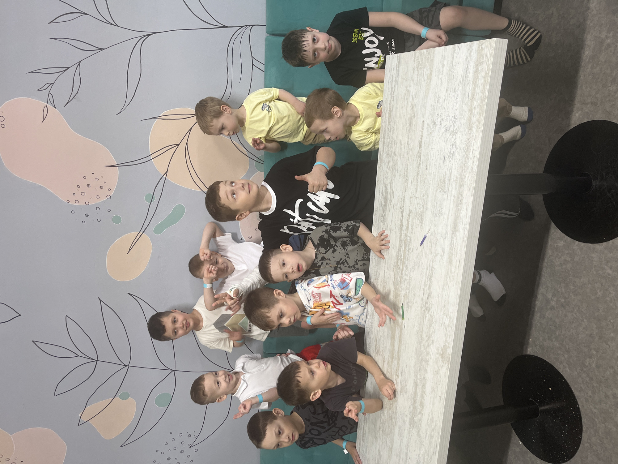 Ten children are gathered around a light-colored rectangular table in what appears to be an indoor recreational or dining area. The scene is in Ulyanovsk, Russia. The children are wearing a variety of casual clothing, including t-shirts and shorts. Some are sitting on a green upholstered bench along a wall painted with a modern design of leaves and abstract shapes. Two black table bases are visible under the table. One boy has striped socks.