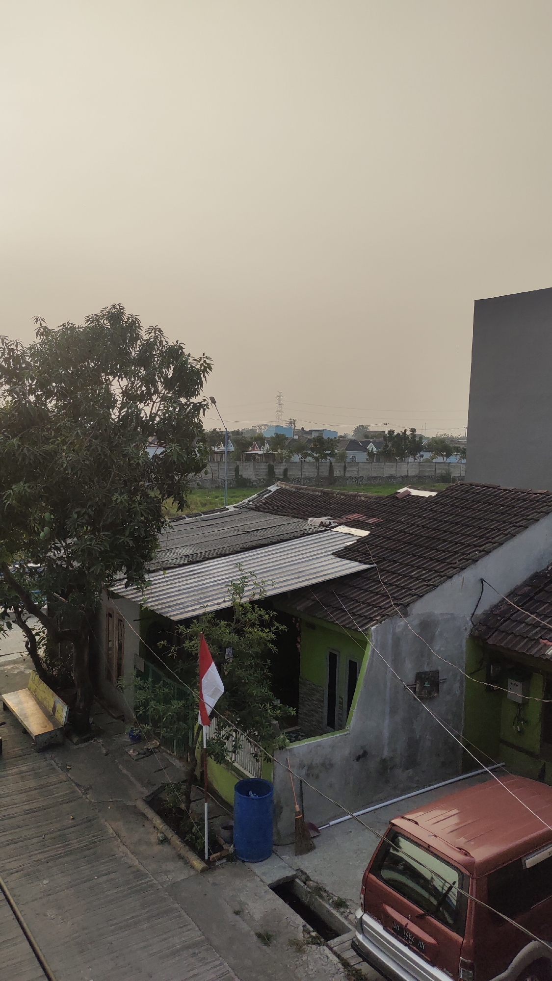 This is a medium-angle, eye-level shot of a residential area in Jalenjaya, Indonesia. The scene is dominated by houses with tiled roofs and plain walls, some painted green and others grey. A large tree with dense foliage stands to the left of the frame, casting shadows. In front of the tree, a concrete bench is visible. A small Indonesian flag on a pole is planted in front of one of the houses, which has a green exterior. Adjacent to the flag is a large blue plastic barrel and a broom leaning against the wall. A red van is parked in the lower right corner of the image, its rear facing the viewer. The ground is paved with concrete, and a small drainage ditch runs along the side of the road. In the background, more houses and some power lines can be seen, with a hazy sky overhead, suggesting it might be late afternoon or early morning. The overall impression is one of a quiet, everyday suburban street scene.