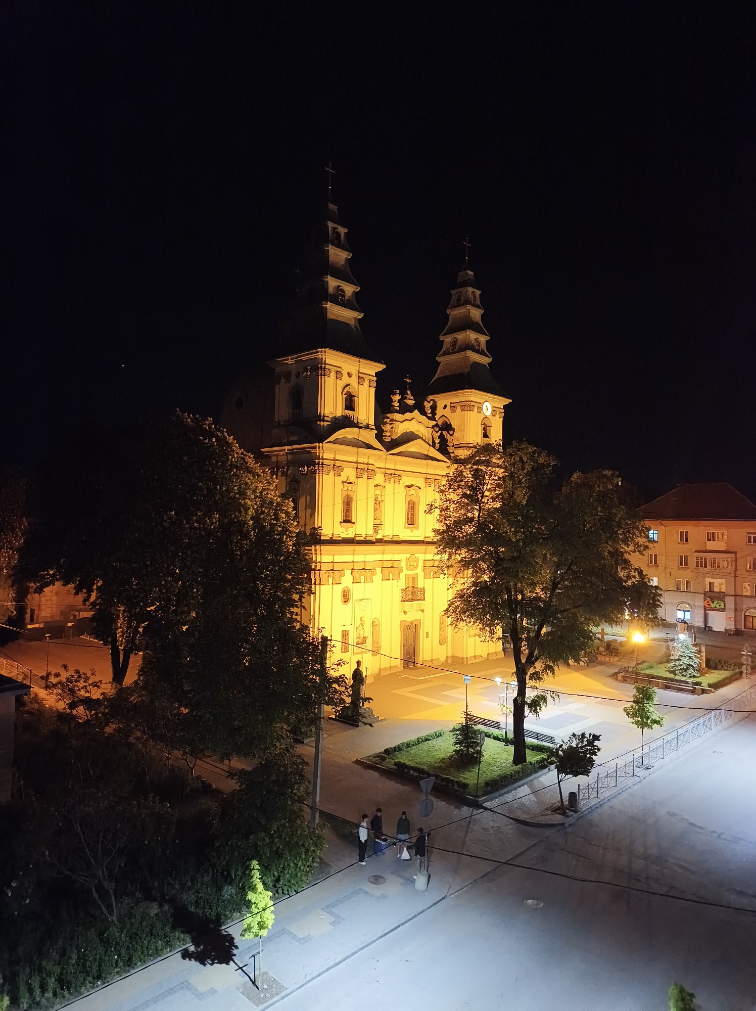 The image shows the Dominican Church in Ternopil, Ukraine, at night. The church is brightly lit, revealing its baroque architecture, including two tall towers topped with crosses. The surrounding area is illuminated by streetlights, and the street appears damp. Several people are standing on the pavement, near the edge of the frame, appearing to converse. Trees surround the immediate area in front of the church. A small park area with greenery, benches and trees is across the street from the church. A building is visible to the right with several windows. The sky is completely dark.