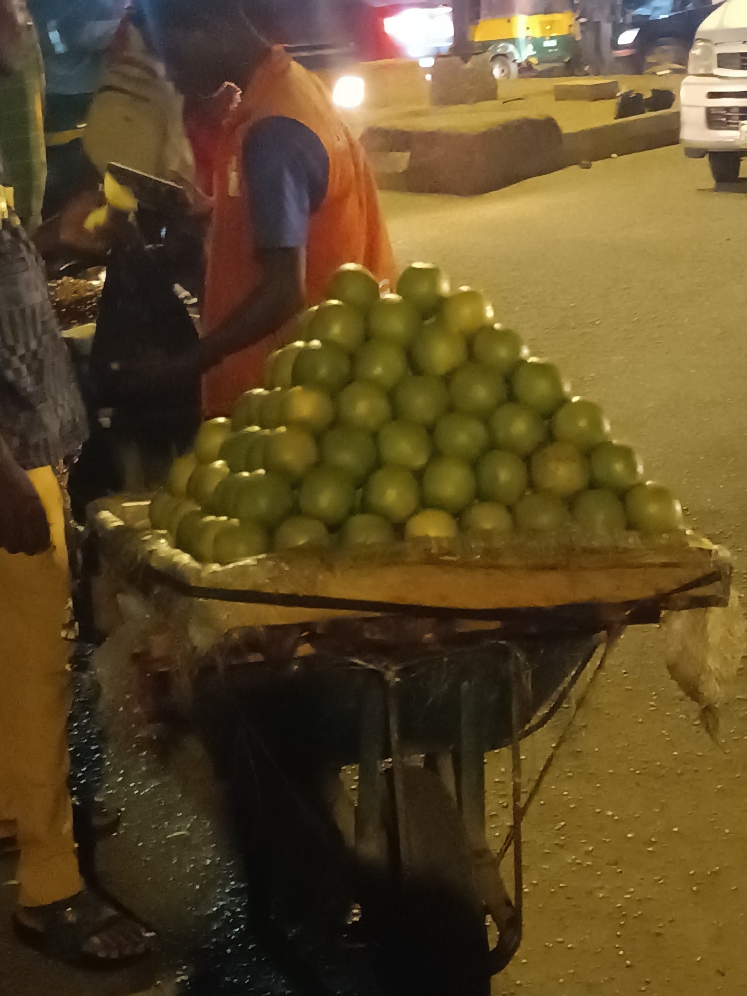 A street vendor in Bukuru, Nigeria, is selling oranges from a wooden cart. The cart is loaded with a pyramid-shaped arrangement of green-skinned oranges. The vendor, dressed in an orange vest over a blue shirt, is standing behind the cart. A customer is partially visible to the left, wearing yellow trousers and a dark shirt. In the background, there are other vehicles and blurred figures indicating a busy street scene. The image is taken at night, and the street is illuminated with artificial lights.