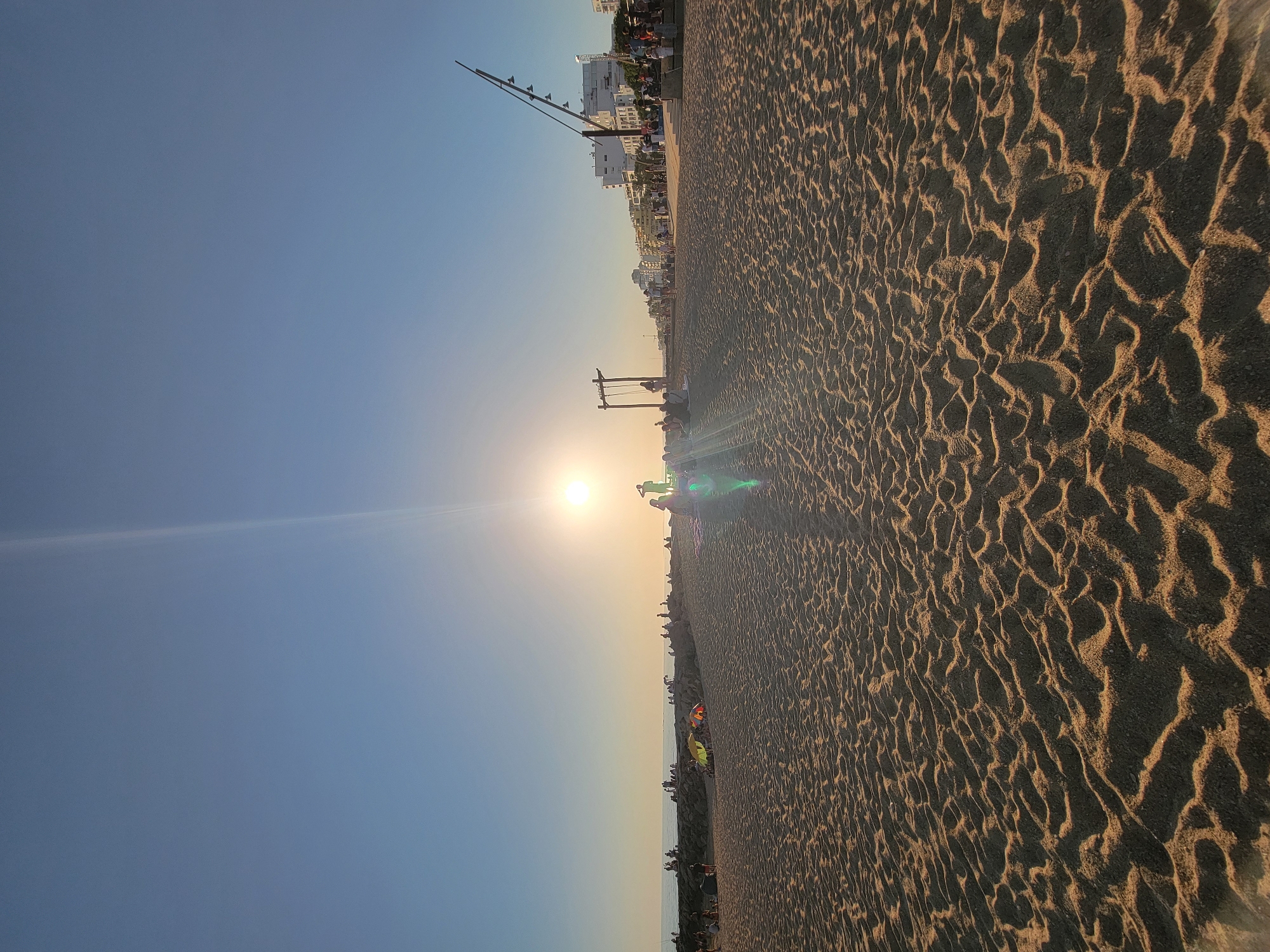The image depicts a sandy beach in Quarteira, Portugal, during what appears to be sunset or sunrise. The foreground is dominated by the textured surface of the sand, characterized by rippled patterns and shadows. In the middle ground, a clear sky transitions to a bright sun, casting a strong glare. Several people are visible on the beach, some sitting, others standing. Towards the right, a series of buildings and a lamppost structure are present, suggesting an urban or developed area adjacent to the beach. In the distance, the sea is visible. A wooden swing set structure is also present on the beach, with at least one person near it. The overall atmosphere is serene and captures a moment of leisure and natural beauty.
