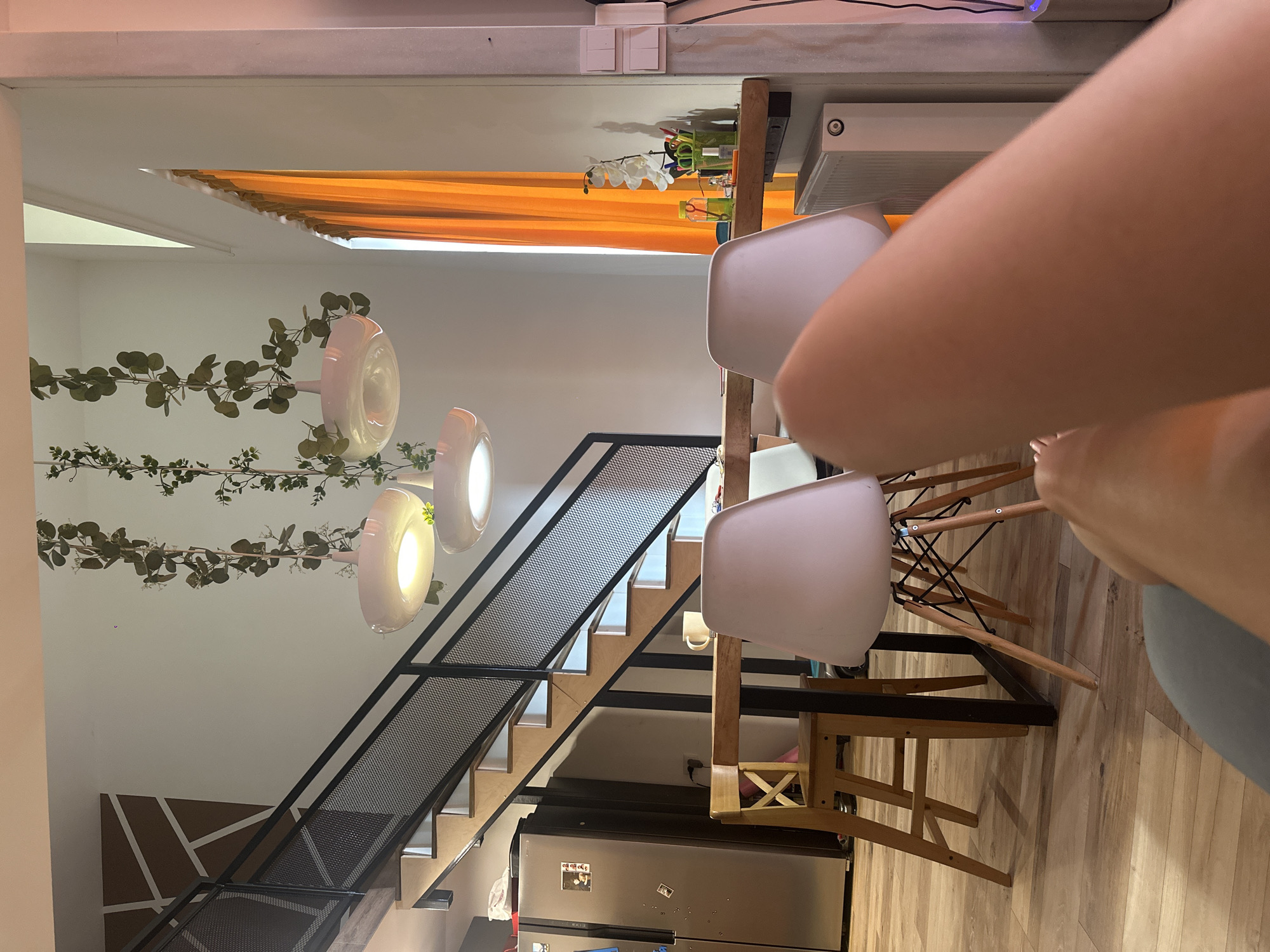 The image shows an indoor scene, likely a dining or living area. The focal point is a light wooden table with several white modern chairs, one of which is partially occupied by a person's arm and leg, suggesting they are seated at the table. The scene has a modern aesthetic, with a minimalist design.
Behind the table, a staircase with wooden steps and black metal railings is visible, leading upwards. Decorative green vines with leaves are draped along the wall near the ceiling, adding a touch of nature to the interior. Three circular pendant lights hang from the ceiling, casting a warm glow.
To the left of the stairs, orange curtains are drawn, hinting at a window or opening. A stainless steel refrigerator is situated at the bottom of the image, with some items, possibly magnets or photos, affixed to its surface. The floor appears to be made of light-colored wooden planks.
The lighting suggests it might be daytime, or the interior lights are on. There is no visible text in the image. The overall atmosphere is relaxed and domestic.