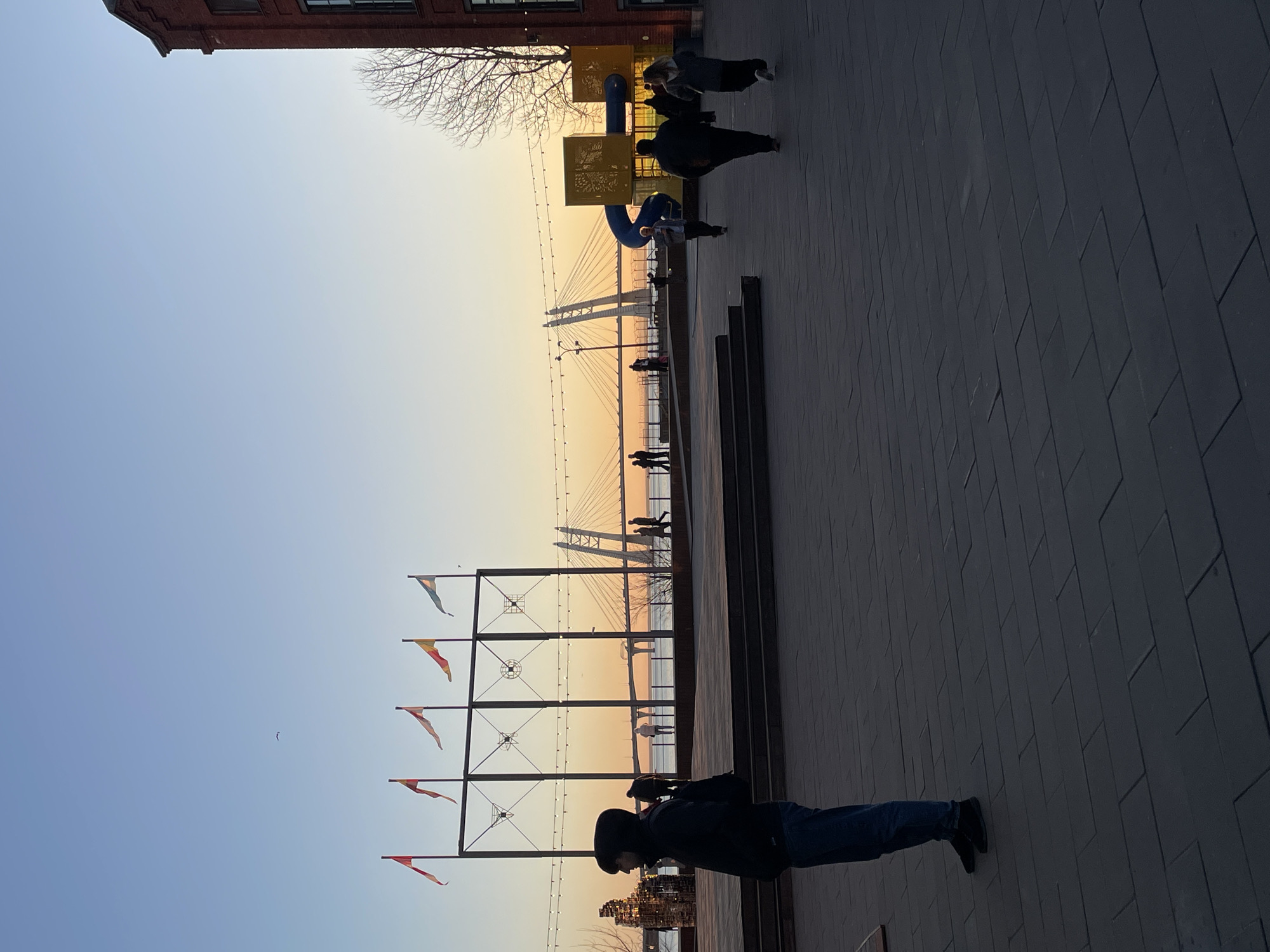 The image shows a promenade in Saint Petersburg, Russia, during a sunset. The prominent feature in the background is the cable-stayed bridge across the Neva River, known as the Western High-Speed Diameter (WHSD) bridge. Along the promenade, there are metal flagpoles with colorful flags. There are people walking along the promenade. A person wearing a black jacket and jeans is standing near the right edge of the image. There are metal structures on the right side. The ground is paved with grey rectangular stones. To the left, there is a playground structure in yellow and blue. The sky is a gradient from blue to orange as the sun sets. In the upper left corner, there is a building with red brick facade.
