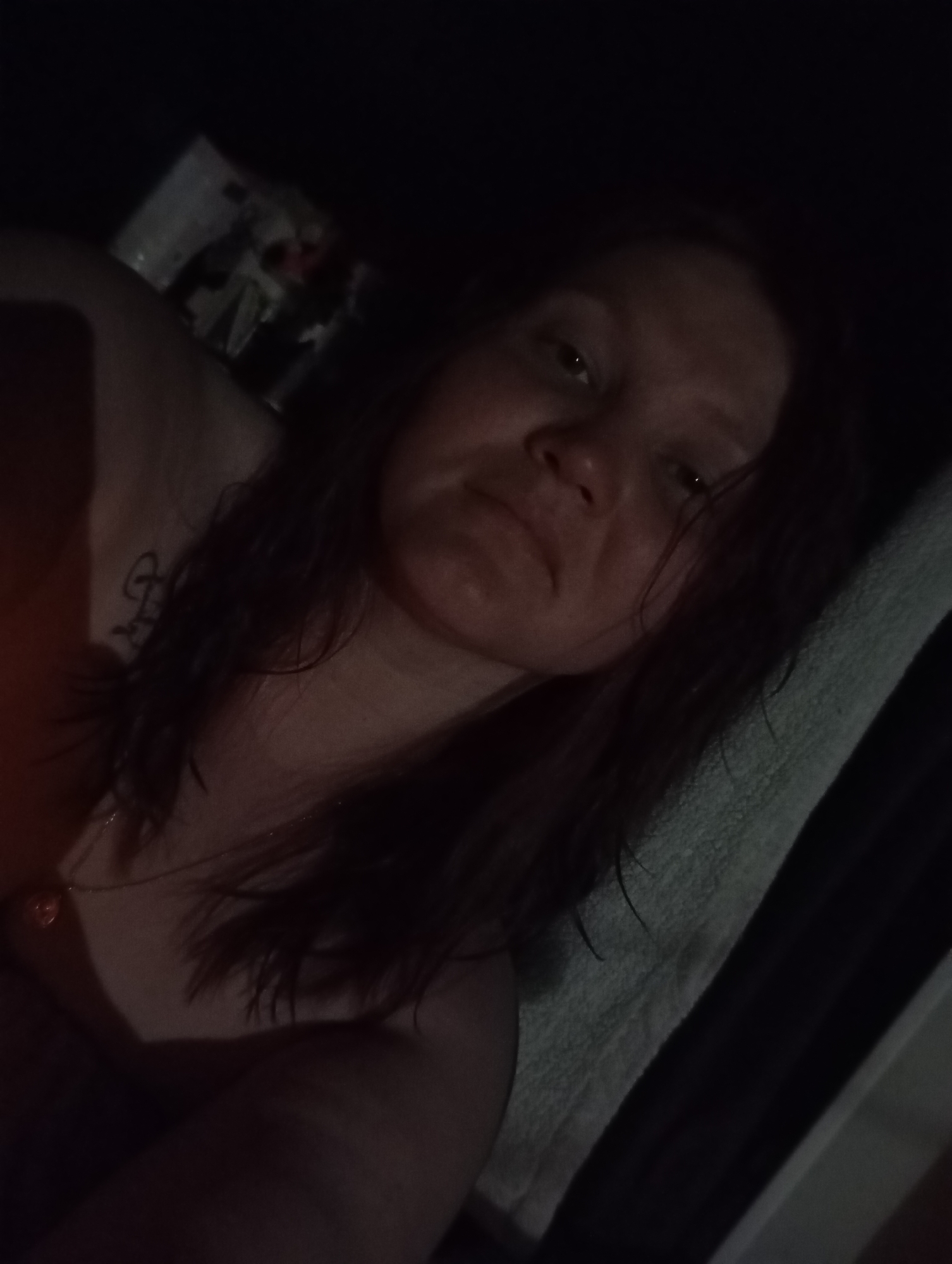 A woman with shoulder-length dark hair is lying on a light-colored pillow or surface. She is facing the camera with a neutral expression. She has a tattoo on her upper chest. A necklace is visible around her neck. A framed picture is partially visible in the background. The location is Unknown, Unknown. The lighting is dim, making details difficult to discern.