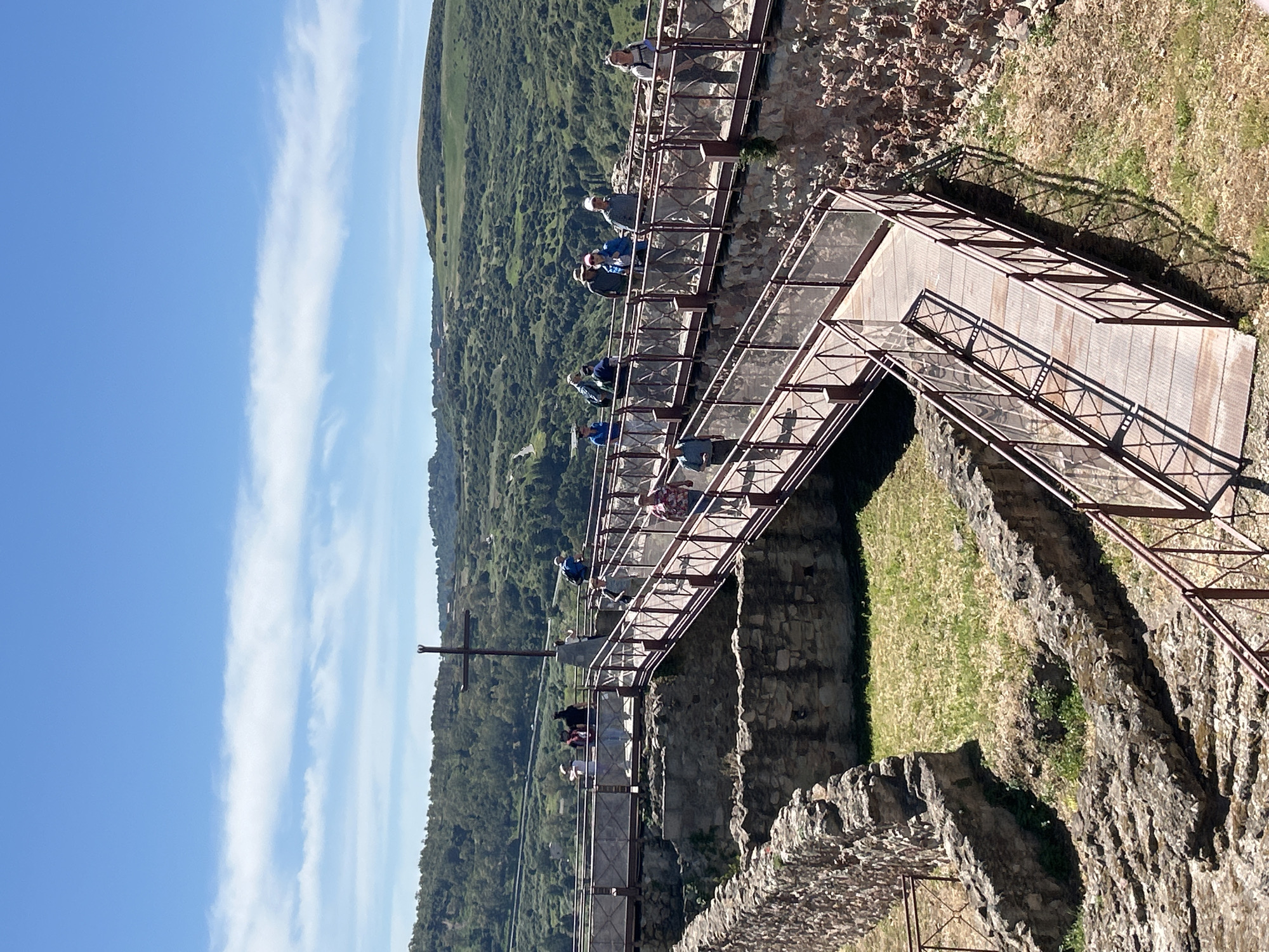 The image shows a metal and wood walkway suspended along a stone wall, likely part of a castle or fortress ruin in Bosa, Italy. A group of people are walking along the walkway, enjoying the views of the surrounding landscape. The landscape includes green hills and valleys under a bright blue sky with some white clouds. In the background, a dark metal cross stands against the green hillside. The construction of the walkway suggests it is a recent addition to allow visitors safe access to the historic site. The sun is shining, indicating it is likely daytime.