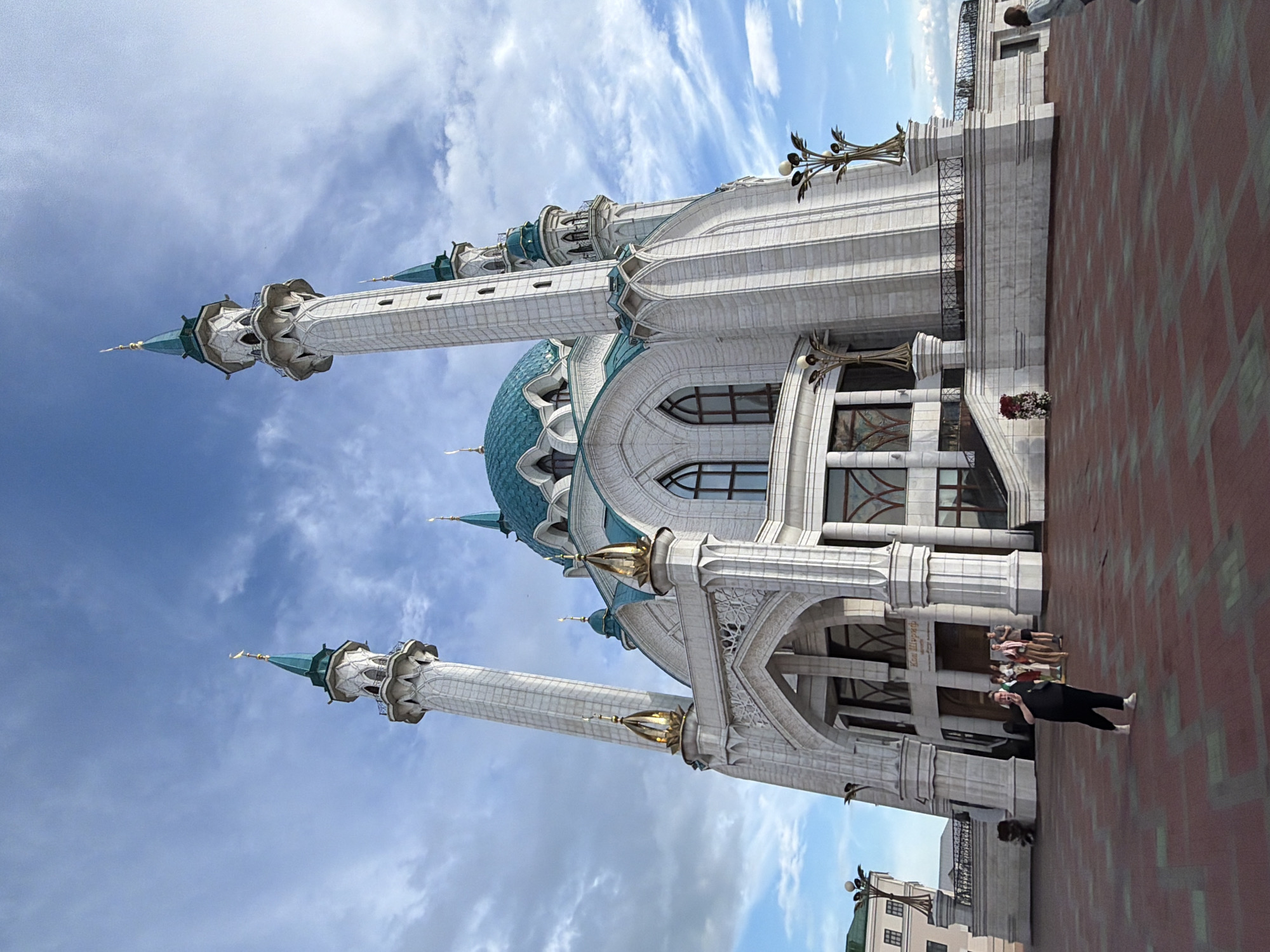 This is a photograph of the Kul Sharif Mosque in Kazan, Russia, taken on a bright, partly cloudy day. The mosque, a prominent landmark, is depicted from a low angle, emphasizing its grand architecture. Two tall, slender minarets with teal-colored spires flank the main building, which features ornate white stonework and turquoise domes. The exterior walls are adorned with intricate carvings and arched windows.
In the foreground, a red brick plaza extends from the mosque. On the plaza, a woman wearing a black top and pants is standing and looking at her phone. Behind her, near the entrance of the mosque, a group of people are gathered. The sky above is a vibrant blue, with wispy white clouds scattered across it, suggesting it's midday. The overall scene conveys a sense of architectural grandeur and a peaceful atmosphere. There is no visible text in the image.