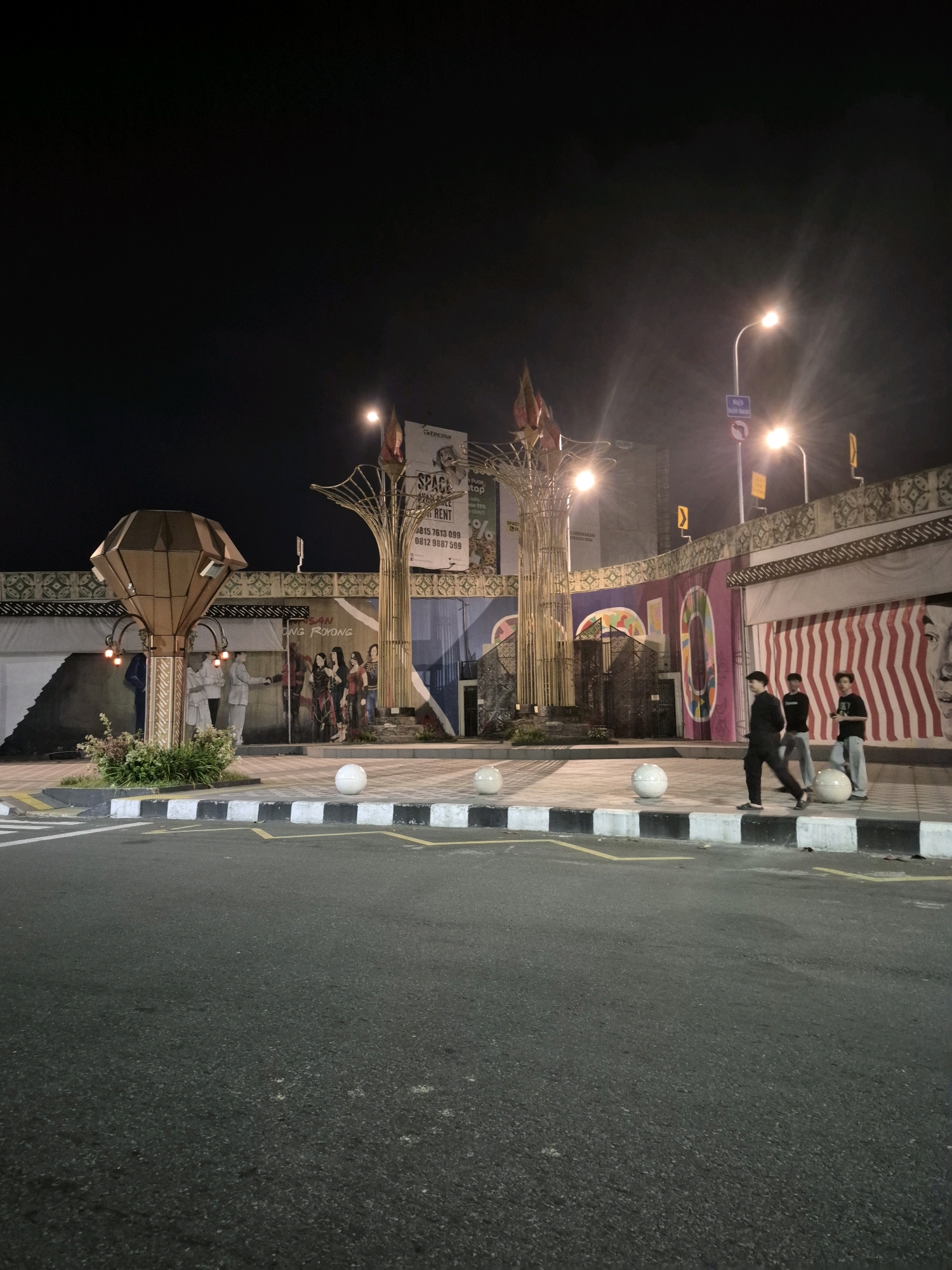 The image captures an outdoor scene at night in Surakarta, Indonesia, likely in an urban or commercial area. In the foreground, a dark asphalt road with yellow lane markings is visible. Adjacent to the road is a sidewalk bordered by a curb with alternating black and white segments. Along the sidewalk, three white spherical objects are placed.
Further back, a decorated pedestrian walkway or plaza is illuminated by streetlights and architectural lighting. On the left, a distinctive diamond-shaped structure is illuminated by red lights at its base. Behind this structure, a mural depicts several figures in what appears to be traditional attire. To the right of the mural, a large, stylized tree-like sculpture with golden elements stands. Above the walkway, a billboard advertises "SPACE RENT" with contact information.
Several individuals are present in the scene. Three young men are walking along the sidewalk. On the left side, near the diamond structure, two figures in white attire are interacting, seemingly part of the mural or a live display.
The background features a building with decorative patterns on its upper facade and red and white striped awnings or panels. Streetlights cast a bright glow, creating lens flares in the image. The overall atmosphere is one of nighttime urban activity, with decorative elements and public art visible.