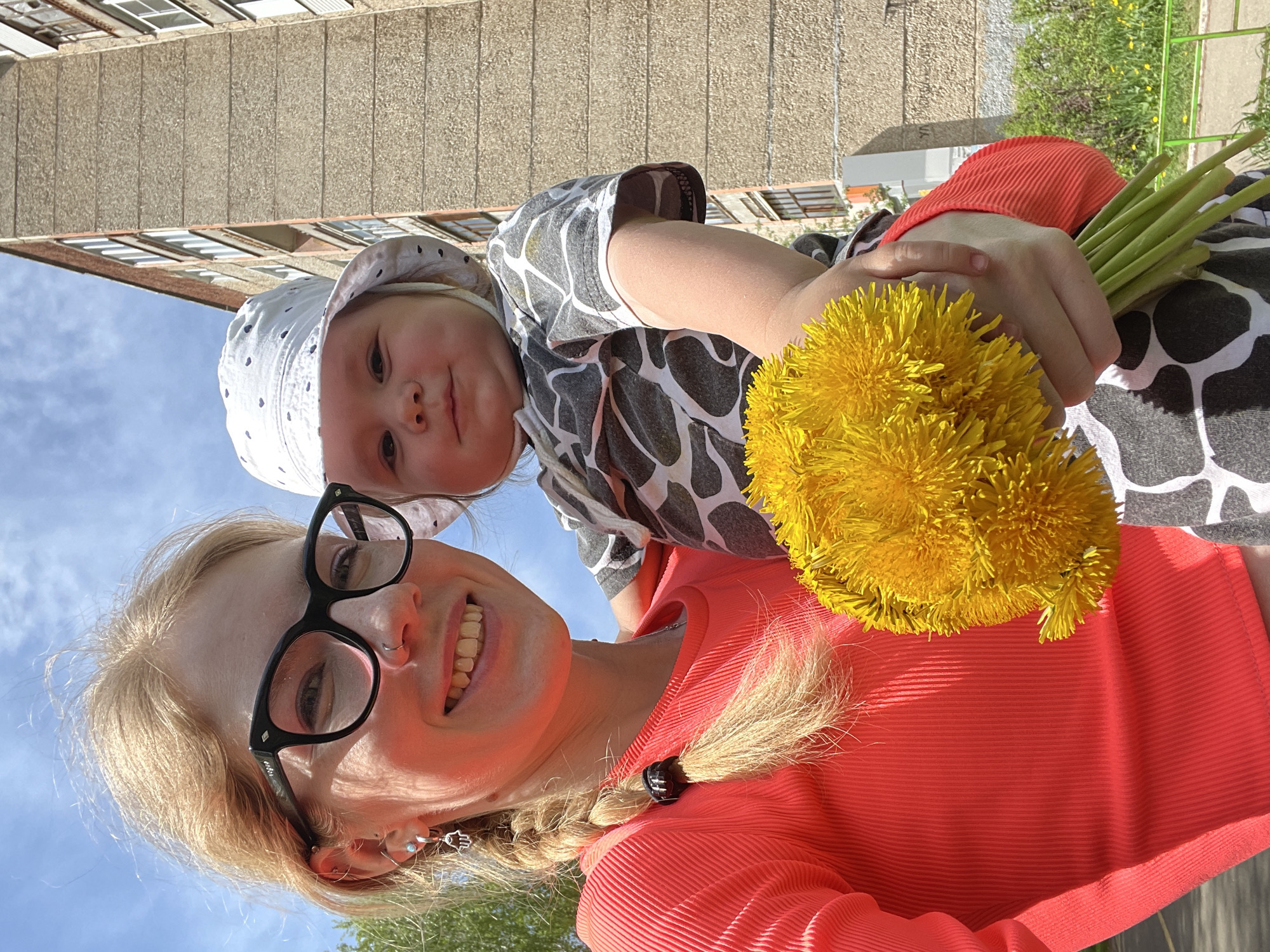 A photograph shows a woman holding a baby while standing outside on a sunny day. The image seems to have been taken from a low angle, looking upwards.
The woman has blonde hair, which is braided on one side and wears glasses. She is smiling and wearing a bright orange long-sleeve shirt. The baby is wearing a hat and a patterned shirt. The woman is holding a large bunch of dandelions in her arms. The buildings and greenery in the background suggest an urban environment. Based on the location request, this is Izhevsk, Russia.
