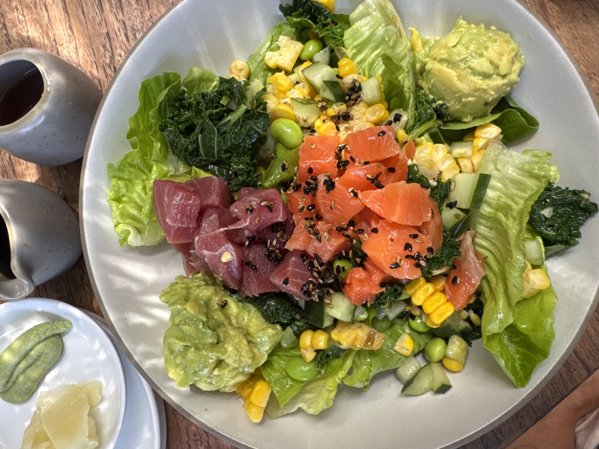 A vibrant poke bowl or deconstructed sushi salad is the central focus of this image, presented in a large, light-colored ceramic bowl on a wooden table. The bowl is generously filled with fresh ingredients arranged in distinct sections.
The base of the bowl features crisp green lettuce leaves. Layered on top are two prominent piles of diced raw fish: one section of dark red, cubed tuna or ahi, and another section of bright orange, cubed salmon. Both fish varieties are sprinkled with black sesame seeds.
Other colorful ingredients are interspersed throughout the bowl: bright yellow corn kernels, light green edamame beans, and diced cucumber. There are also portions of a darker green leafy vegetable, possibly kale or cooked spinach, and two scoops of mashed avocado or guacamole, one at the top right and another at the bottom left of the bowl.
To the left of the main bowl, two small, grey ceramic pitchers, likely containing soy sauce or dressing, sit on the wooden table. Below them, a small white ceramic plate holds accompanying condiments: a dollop of green paste (most likely wasabi) and a pile of thinly sliced pickled ginger.
The overall scene depicts a fresh and appealing meal, set on a warm-toned wooden surface, suggesting an indoor dining environment. No people are visible, and there is no visible text in the image.