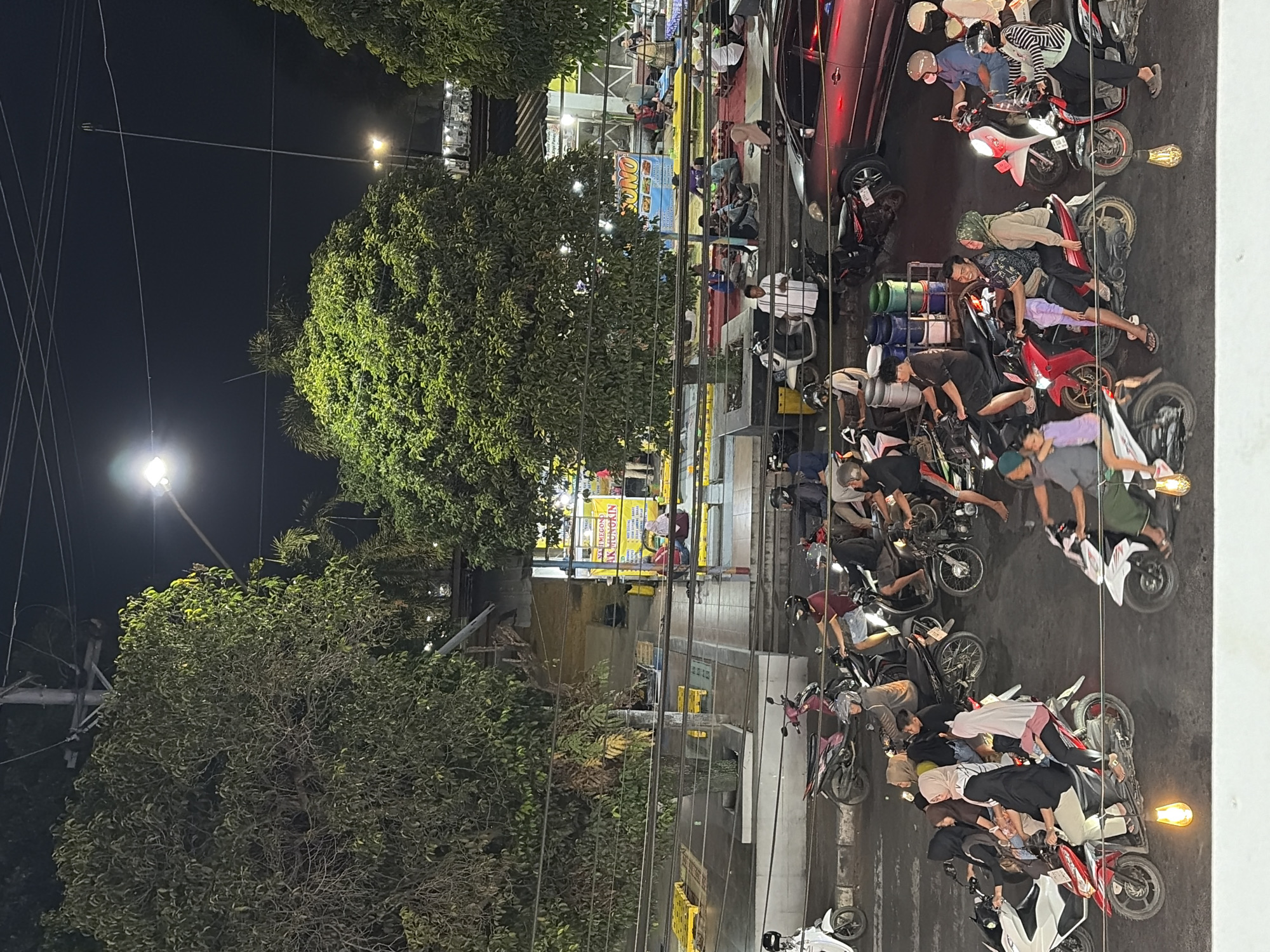 The image captures a nighttime scene in Batang, Indonesia, likely an outdoor seating area or market. Numerous people are present, many of them on or around motorbikes. Several motorbikes are parked or in motion, some carrying multiple passengers. People are dressed in casual clothing, with many women wearing hijabs. Some individuals are seated on the ground, while others are standing. The environment includes trees, streetlights, and buildings in the background, suggesting a busy urban area. Electrical wires are visible overhead. The overall impression is one of a lively evening gathering or a commercial district with food stalls or shops.