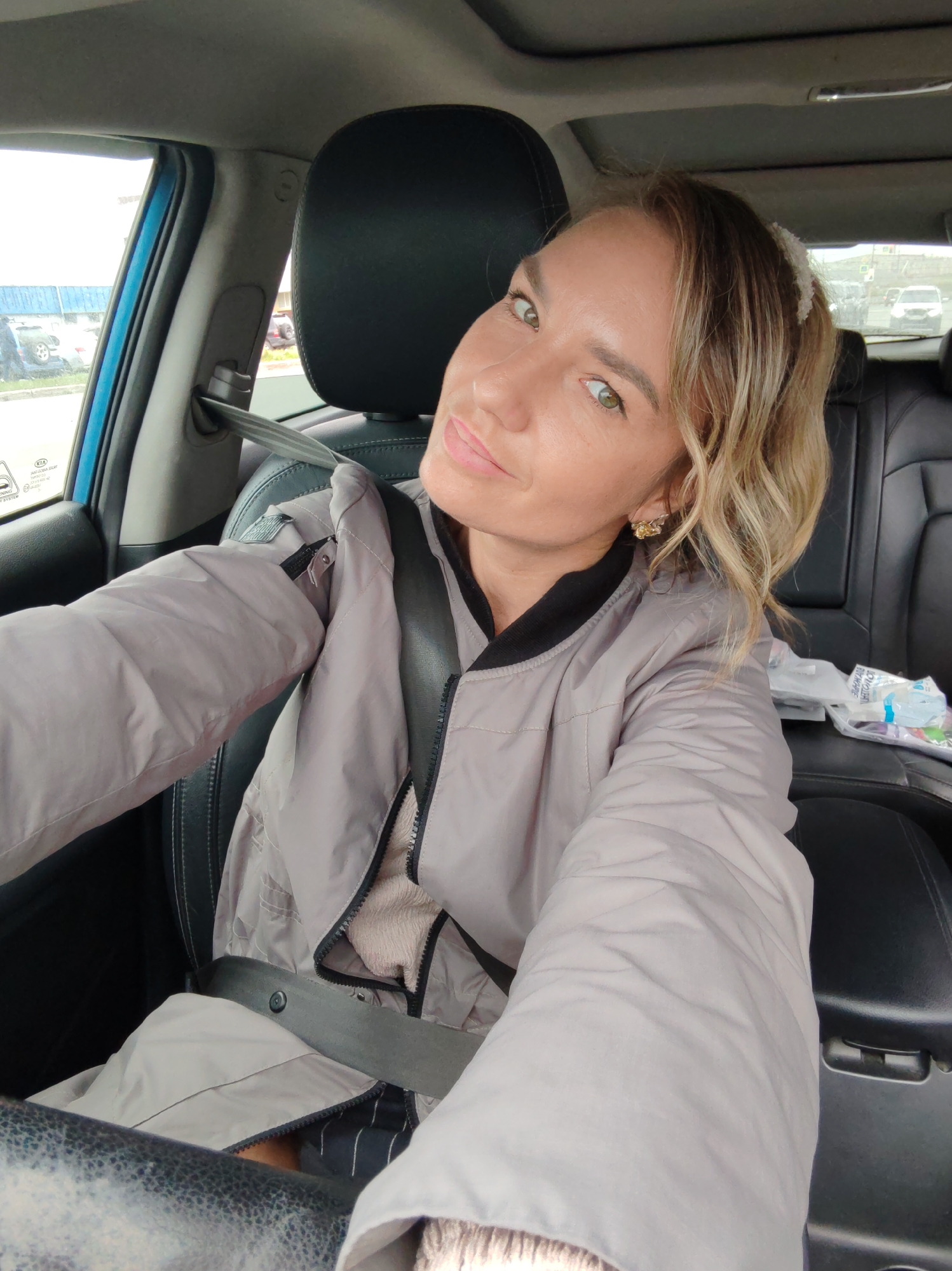 A blonde woman is sitting in the driver's seat of a car, taking a selfie. She is wearing a light gray puffer jacket over a tan sweater and dark striped pants. A seatbelt is fastened across her body. Her hair is partially pulled back with a white clip. She is wearing gold earrings. The interior of the car is visible, including the black leather seats, the steering wheel, and a portion of the dashboard. Outside the car window, other vehicles and a light blue building are visible, suggesting an urban or commercial area. The image was taken in Petropavlovsk-Kamchatsky, Russia.
