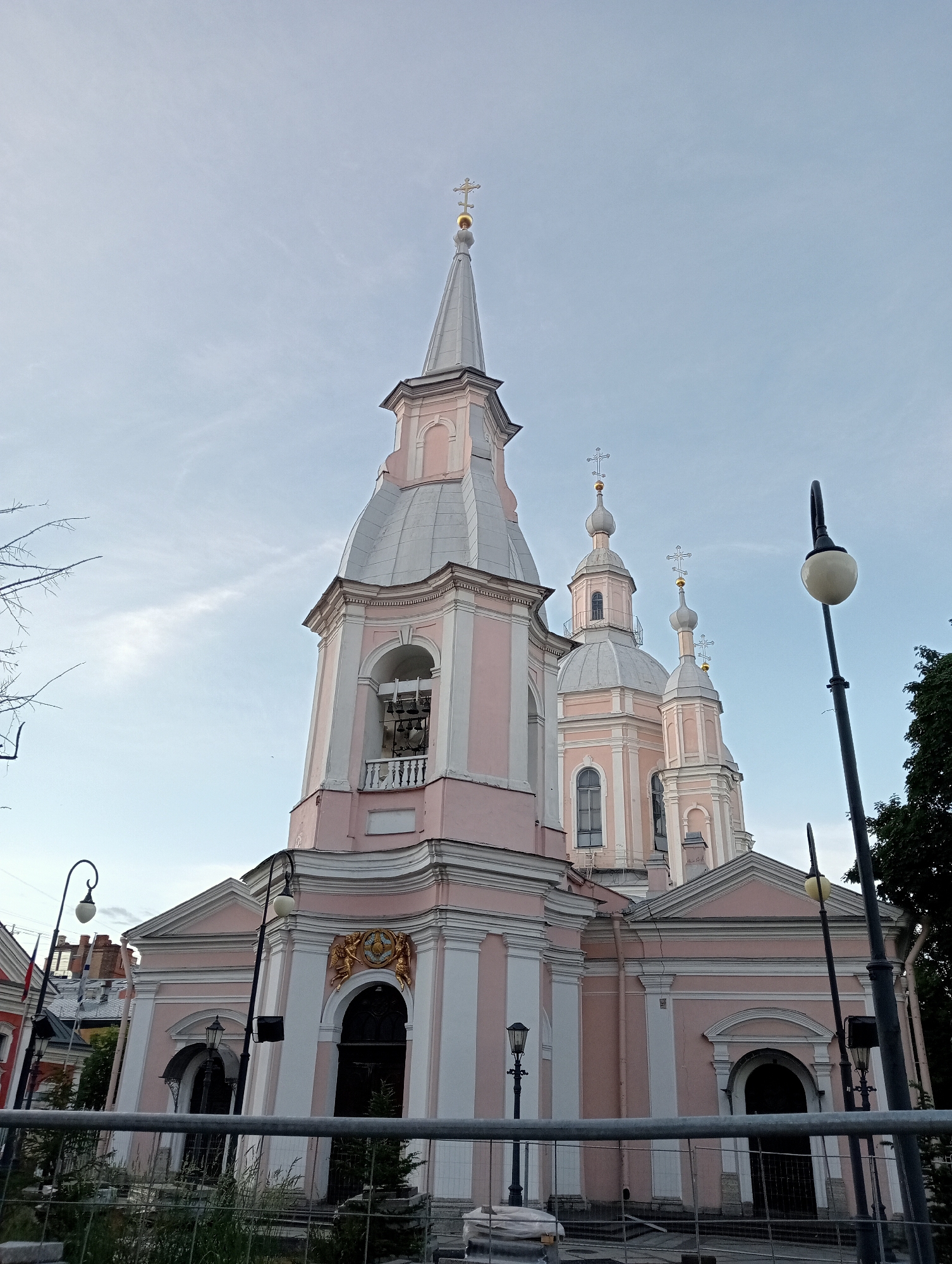 The image shows a low-angle, wide shot of the pink and white Smolny Cathedral in Saint Petersburg, Russia, under a pale blue, cloudy sky. The cathedral features multiple domes and spires topped with gold crosses. A bell tower is visible on the left side of the main structure, with a balcony containing several bells. Several ornate architectural elements, including arched doorways and decorative moldings, are visible on the facade. A tall black lamppost with a white globe is positioned to the right of the cathedral, and another smaller, black lamppost is visible on the left. A metal fence runs across the foreground of the image, separating the viewer from the cathedral grounds, which are partially obscured by greenery and construction materials. The overall scene depicts a quiet, perhaps late afternoon or early morning, view of the historic landmark.