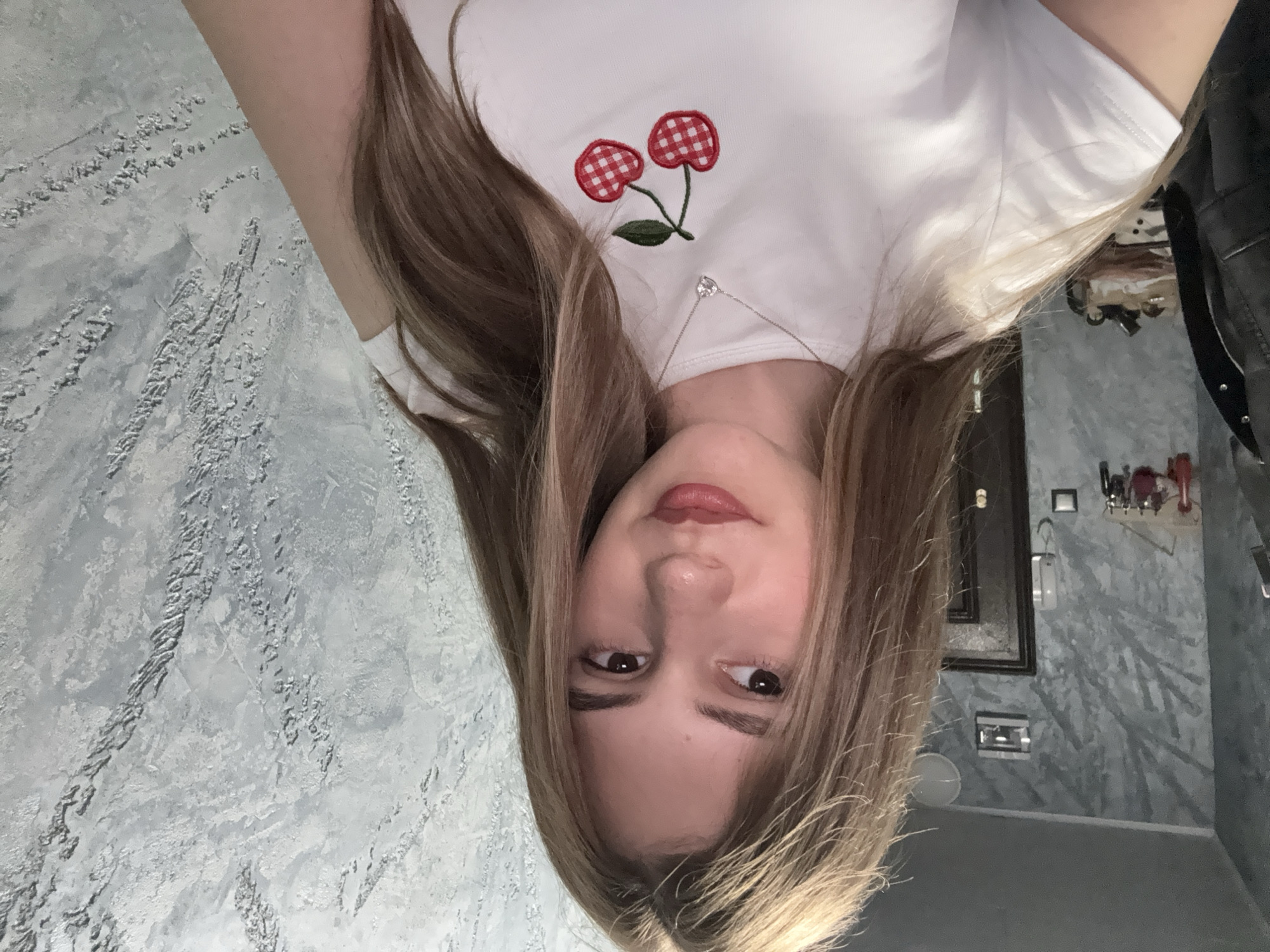 A young fair-skinned woman with long, light brown hair is facing the camera. She is wearing a white t-shirt with a red and white checkered cherry design on the upper chest area. A delicate silver necklace with a small pendant is visible around her neck. The background consists of a textured gray wall. A framed picture hangs on the wall. A black leather jacket is visible in the periphery. It is not possible to determine the location in the city of Panagyurishte, Bulgaria from the image alone.