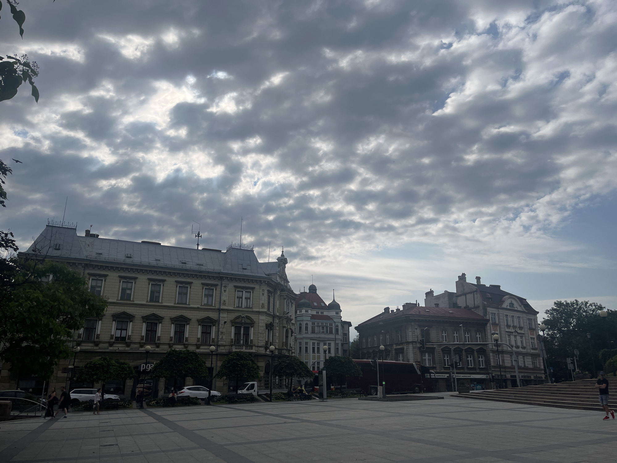 The image shows a wide-angle view of a city square under a cloudy sky. In the foreground, a paved area with a geometric pattern is visible. Several people are walking or standing in the square. On the left side, trees and a dark-colored car are present. The middle ground features a row of buildings with ornate architectural details, including multiple stories, windows, and decorative elements. Some of these buildings appear to house businesses, indicated by visible signage. A large, dark-colored vehicle, possibly a bus or van, is parked in front of one of the buildings. To the right, a set of wide stone stairs leads upwards, with a lone person standing at the top. Trees and more buildings are visible further in the background. The overall impression is of a public space in a European city.