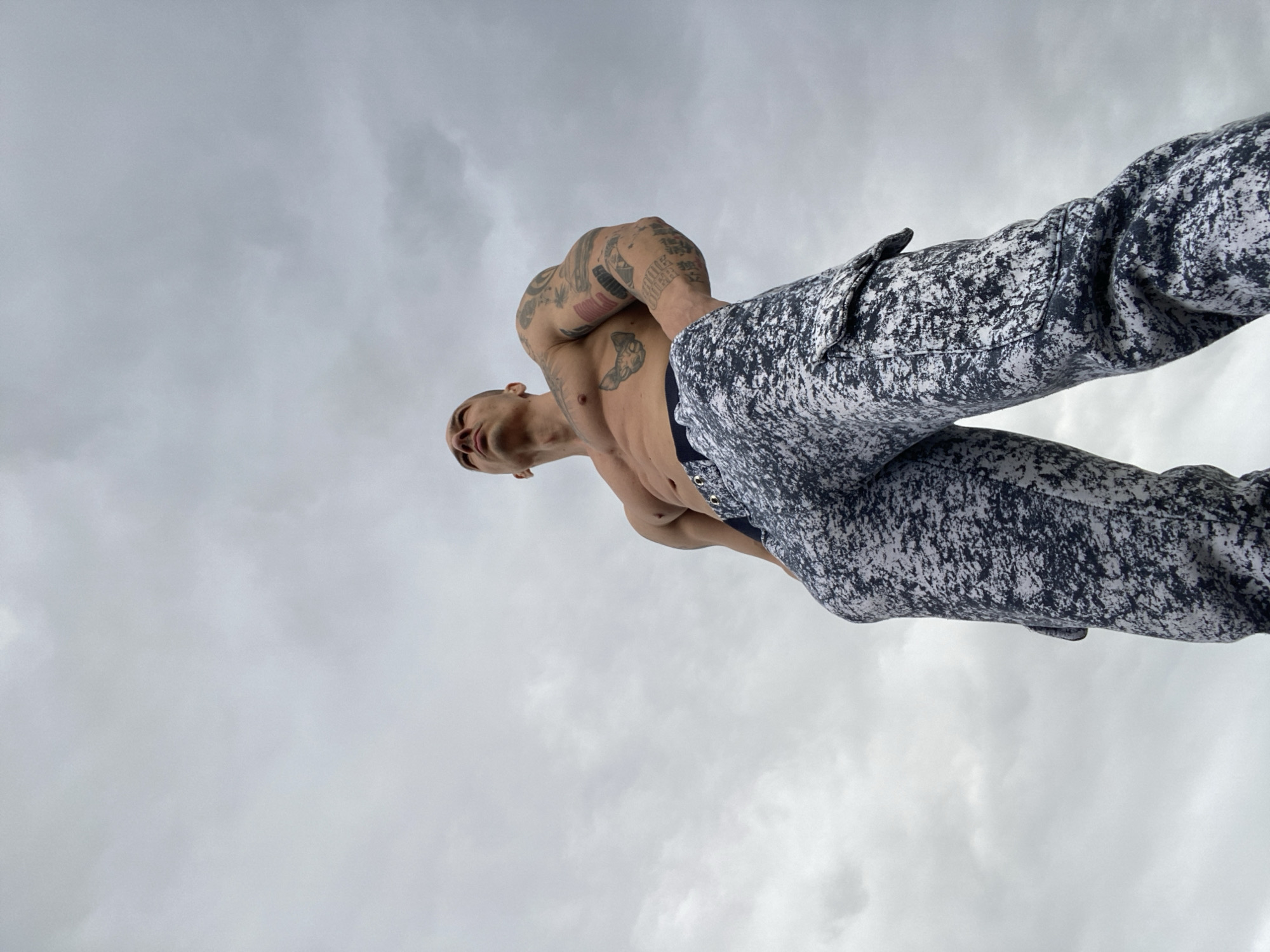 A low-angle, upward-looking photograph captures a man against a cloudy sky. The man is fair-skinned and has a shaved head. He is wearing pants with a black and white textured pattern and is shirtless, revealing his torso. He has tattoos on his arms and chest. The sky is overcast with varying shades of gray clouds filling the frame. The perspective suggests the photo was taken from ground level, looking up at the man. No specific location in Pleven, Bulgaria, can be determined from the provided image.