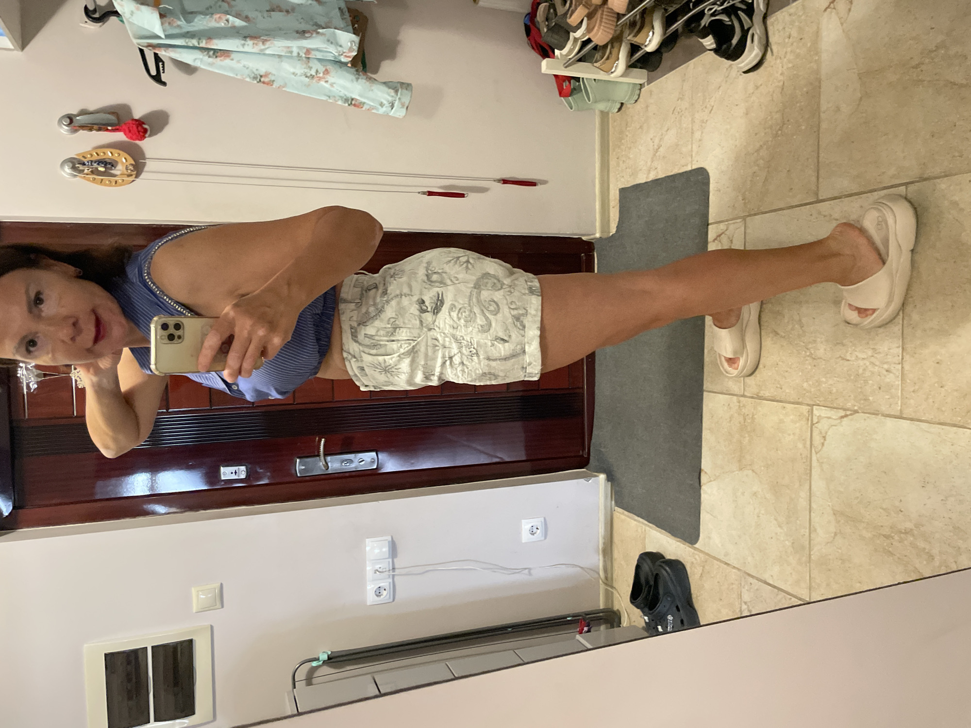 A woman, appearing to be in her 40s or 50s, stands in a hallway, taking a mirror selfie with her smartphone. She is wearing a blue and white striped sleeveless crop top and white shorts with a patterned print. The hallway has light-colored tiled flooring and a wooden door. There are several electrical outlets on the wall, a shoe rack with various shoes, a hanging piece of clothing with a floral pattern, and some decorative items on the wall. The woman's right arm is raised, touching her neck. The overall scene suggests a domestic setting. Based on the presence of a shoe rack with sandals and casual attire, the location is likely a residential apartment. While no explicit visual cues indicate the city of Sochi, Russia, the user's prompt specifies this as the location.