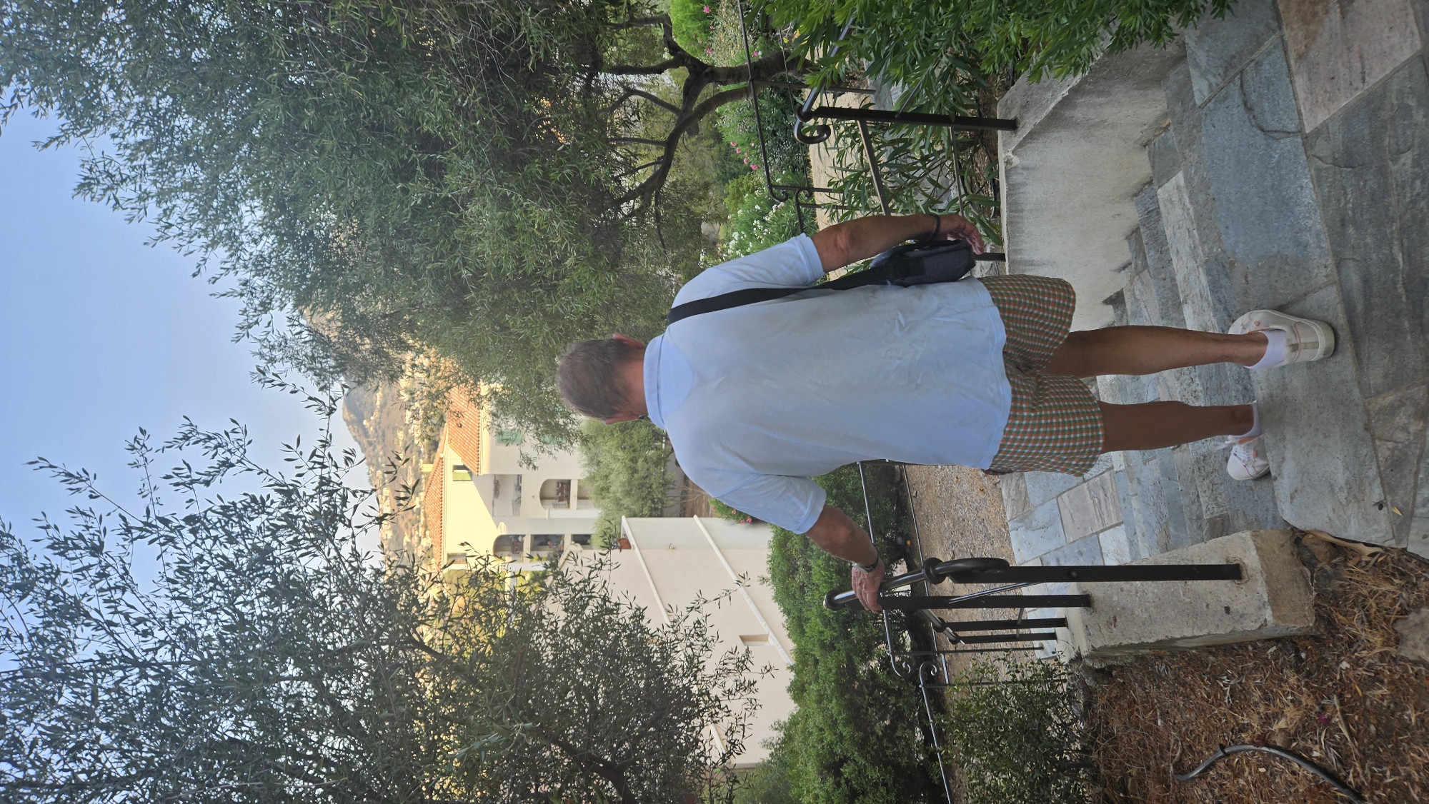 An adult male, viewed from behind, walks down a set of stone stairs outdoors. He is wearing a light blue polo shirt, plaid shorts, and white sneakers. A dark bag is slung over his right shoulder. He is holding onto a black metal railing on his left. The stairs are flanked by greenery and trees. In the background, there are white buildings with terracotta roofs and a hillside. The sky is clear and blue. This scene is located in L'Île-Rousse, France.