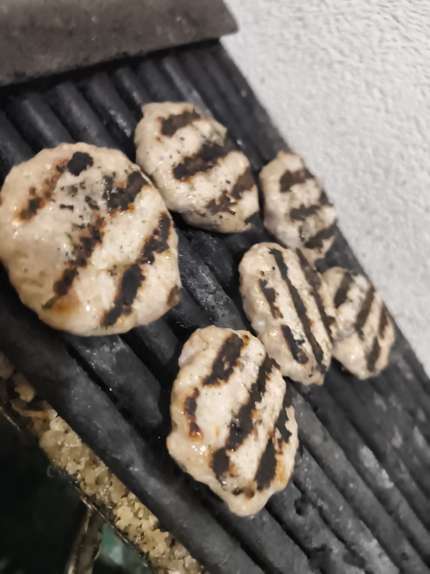 A close-up, angled photograph captures several irregularly shaped, light-colored patties or meatballs being grilled. Approximately seven pieces of food are visible, arranged across dark, ridged grill grates. Each piece displays prominent, charred black grill marks, indicating they have been cooked over high heat. Some patties show faint specks of green, possibly herbs.
The food rests on robust black grill grates, which extend from the lower left to the upper right of the frame. In the upper left, an out-of-focus, dark, flat object is partially visible. The background on the right side of the image consists of a bright, possibly white or off-white, textured surface, resembling a wall or plaster. In the lower left corner, beneath the grill grates, a section of a metal tray or pan is visible, containing a granular, light-colored substance that appears to be cooking residue.
The image depicts the activity of grilling food. There are no people visible, and no discernible text. The lighting is uniform, suggesting either an indoor setting or a well-lit outdoor area. While the context of Plovdiv, Bulgaria, is provided, there are no specific visual cues within the image to confirm this location.