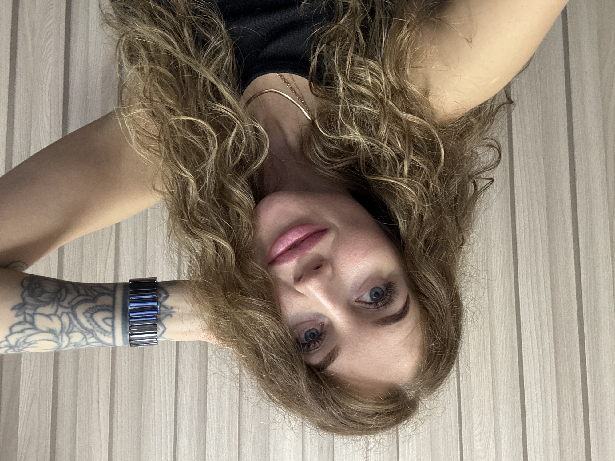 A woman is lying on her back, her head angled towards the camera. She has fair skin, light brown, curly hair, and blue eyes. She is wearing a black top and a gold chain around her neck. Her left arm is raised, with a watch on her wrist and a tattoo visible on her forearm. The background appears to be a vertically striped wall. The location is stated to be Chaikovsky, Russia, although there is nothing in the image itself to confirm this location.