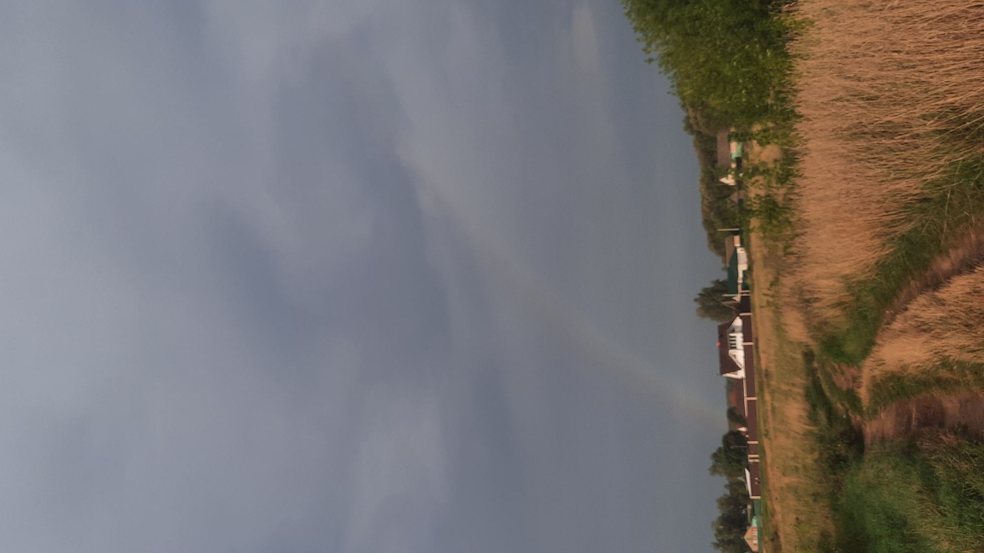 A rural landscape under a cloudy sky features a dirt path winding through dry grass and green vegetation. In the background, a cluster of houses with fences is visible, and a faint rainbow arcs across the sky. The scene appears to be in the vicinity of Shklov, Belarus.