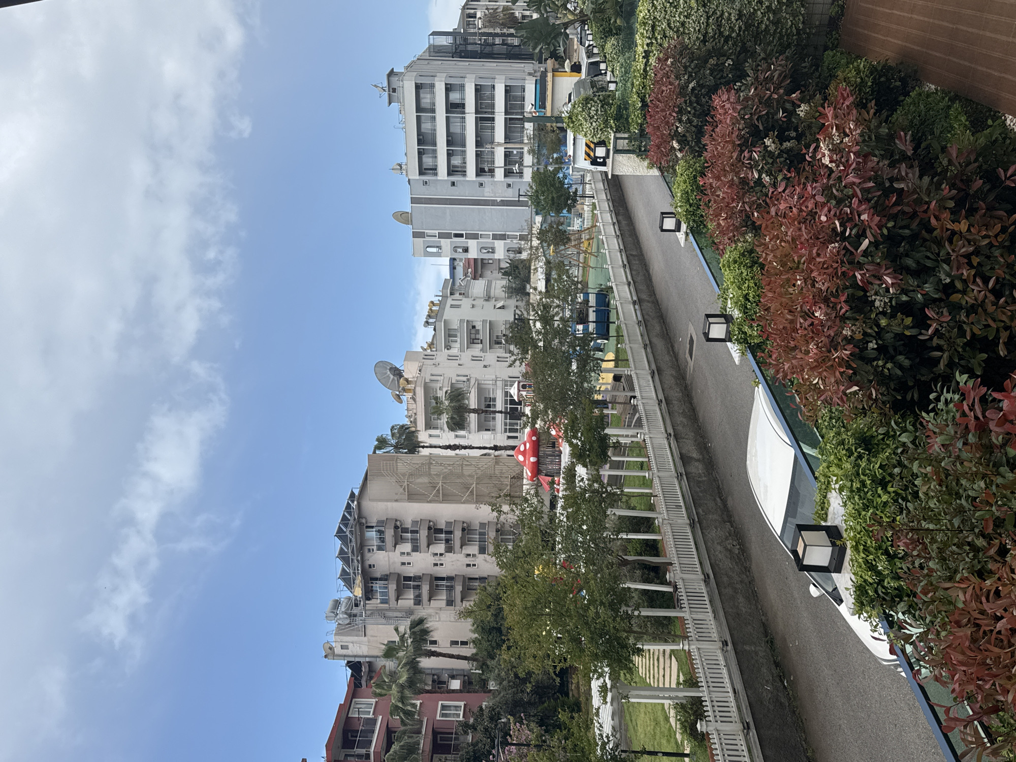 The image shows a street with buildings on one side and vegetation on the other. The buildings are mostly white or light-colored and appear to be residential apartments. There is a playground visible with a red mushroom-shaped structure. The vegetation consists of trees, shrubs, and bushes, some with red leaves. A white fence runs along the edge of the street. The sky is blue with some clouds. The location is an urban area, though the specific city is unknown.