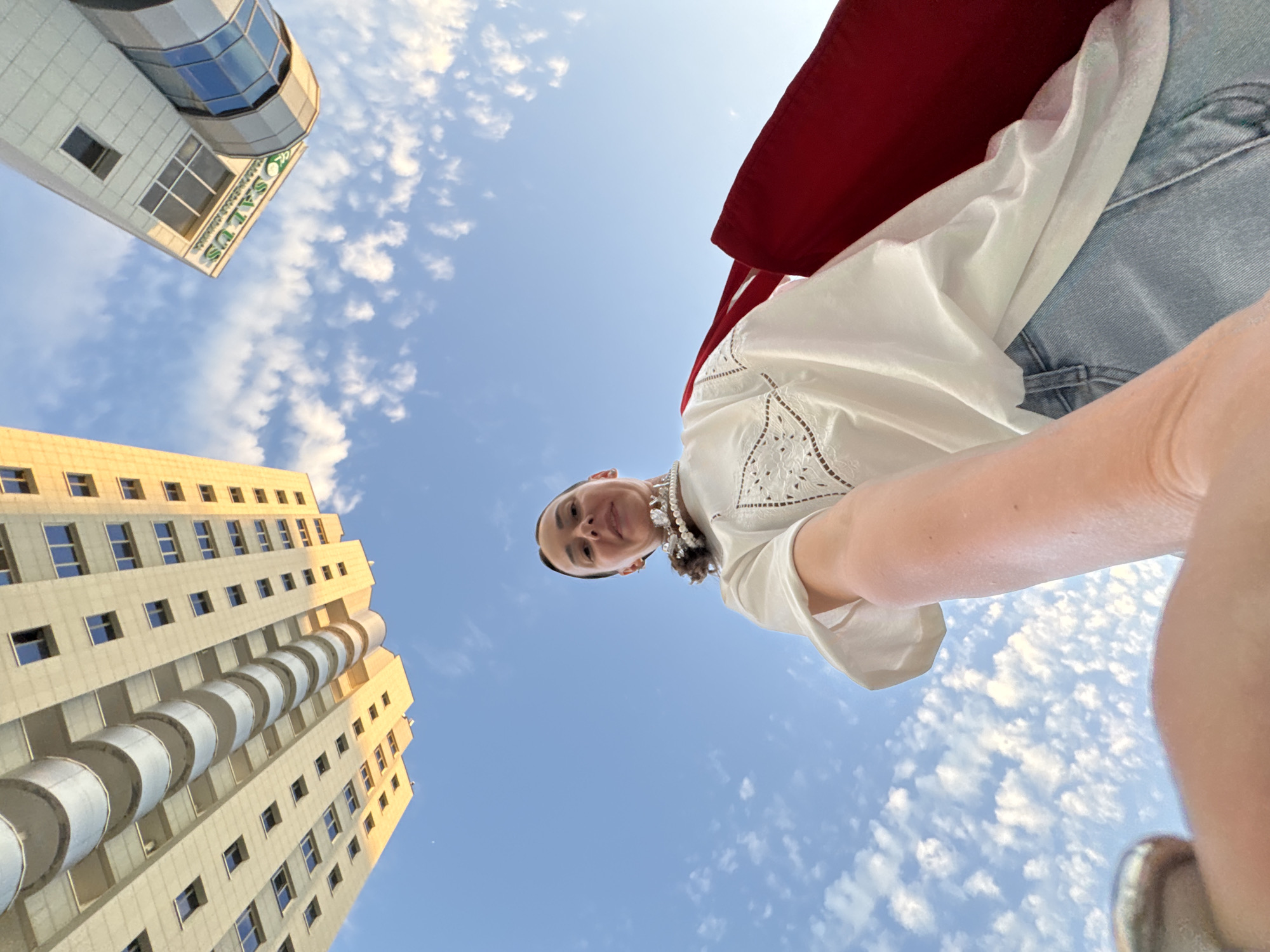 A woman, viewed from a low angle, stands against a blue sky with scattered white clouds. She is wearing a white, loose-fitting top with intricate embroidery along the neckline and shoulders, a pearl necklace, and blue jeans. A large red tote bag is slung over her left shoulder. In the background, two modern buildings are visible, one tall and beige with rounded balconies, and another shorter, white building with a curved glass section and signage that reads "ROSALIA US". The perspective suggests the image was taken in Sochi, Russia.