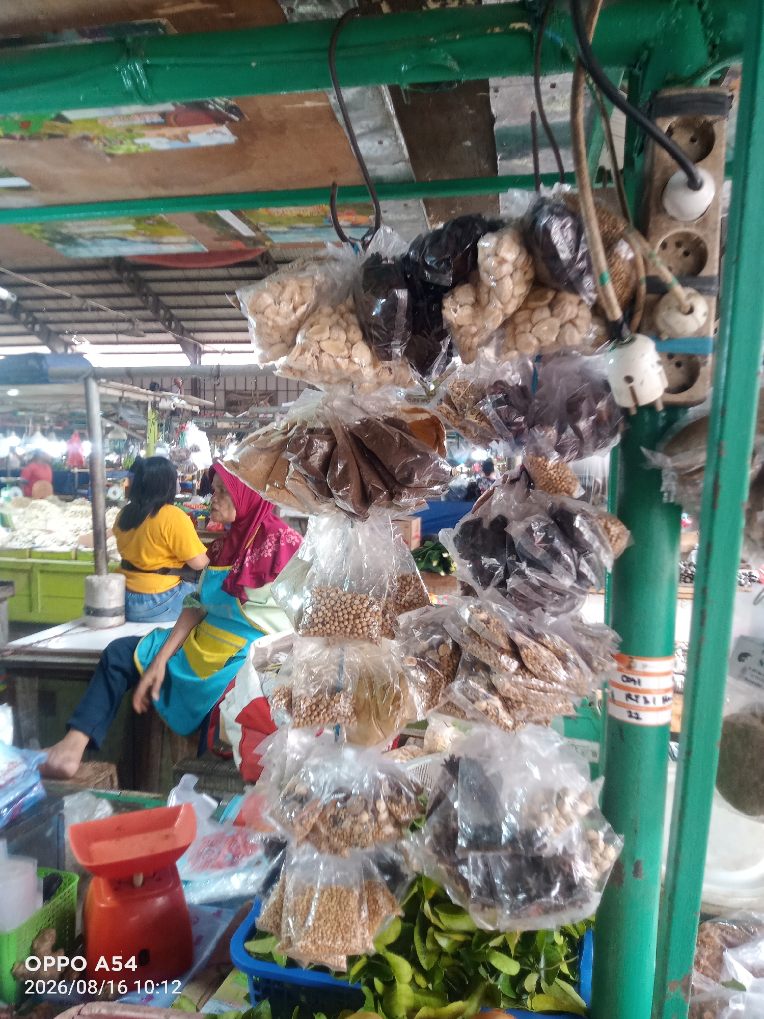 This is a medium shot of a market stall in Jambi, Indonesia, focusing on a display of various goods packaged in clear plastic bags. The goods are suspended from a green metal frame and consist of nuts, seeds, and what appear to be dried herbs or spices. In the background, two women are visible. One, wearing a yellow t-shirt and blue jeans, is sitting with her legs extended. The other, wearing a pink hijab and an apron with yellow and blue accents, is also seated. An orange mechanical scale is visible in the foreground, along with a green plastic basket containing leafy vegetables. The overall environment suggests an outdoor or semi-open market setting.