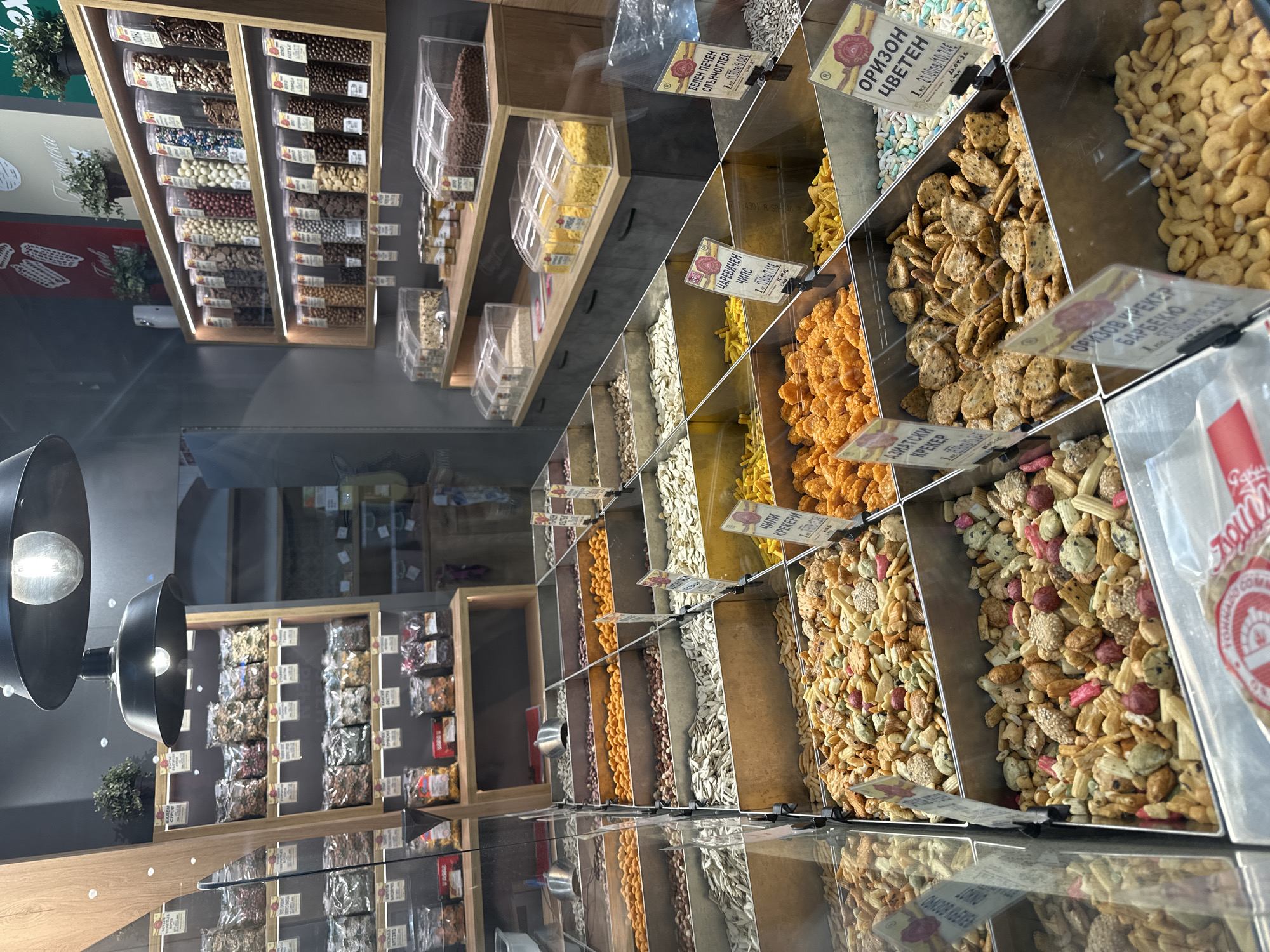 The image shows a view inside a shop specializing in nuts, seeds, and snacks in Varna, Bulgaria. The food items are displayed in bulk containers, each with a small sign denoting the product name and price in Bulgarian and Euro currencies. The snacks vary from sunflower seeds and rice crackers to sweet treats. In the background, there are shelves filled with packaged snacks, as well as bags of nuts. The perspective of the photo is looking down at the shelves, capturing both the depth of the store and the reflections on the glass surfaces. The interior lighting includes spotlight fixtures, with the bulb exposed. The scene suggests a well-stocked and orderly retail environment.