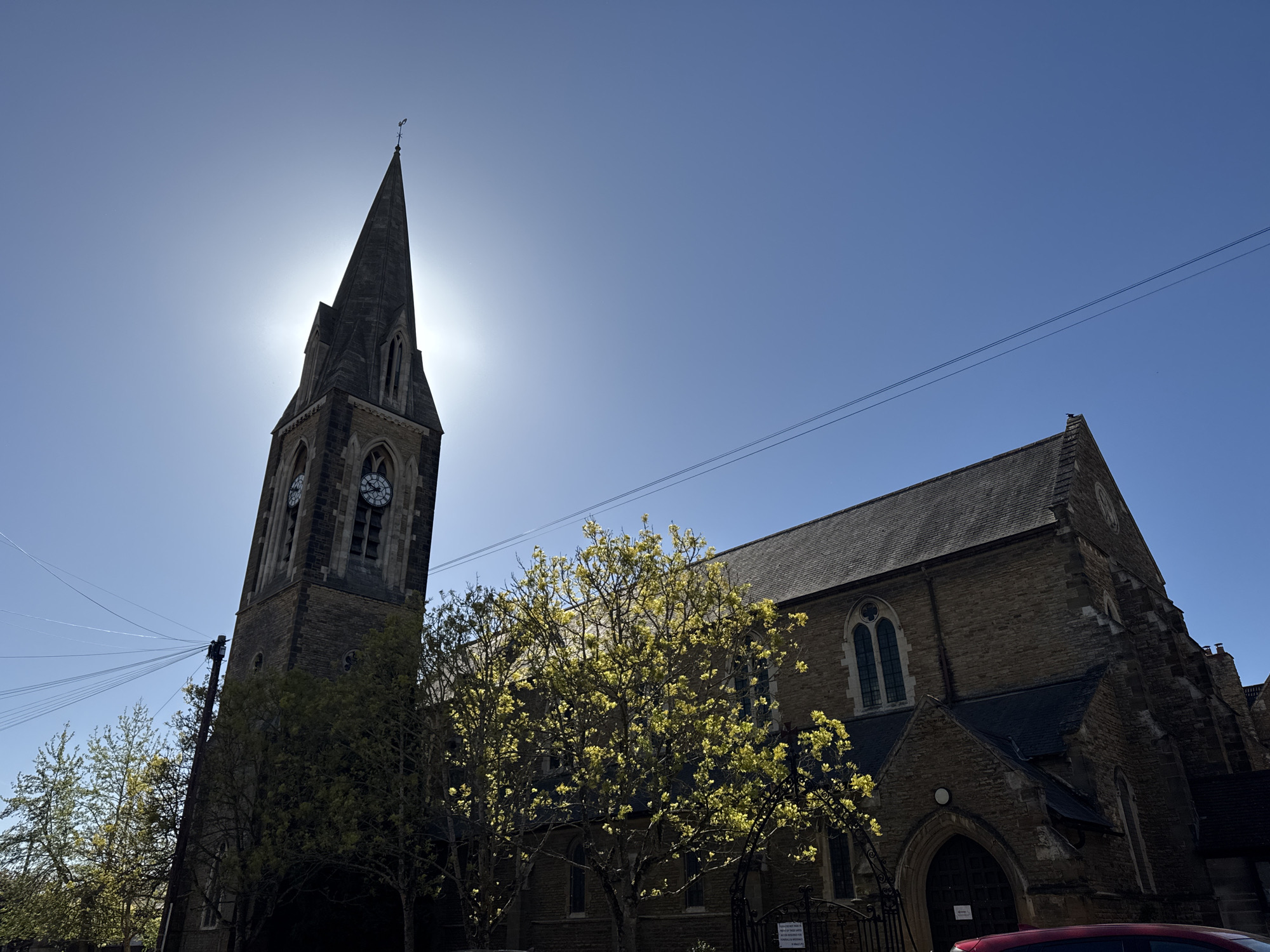 A low-angle, wide shot captures a stone church with a prominent spire under a bright, clear blue sky. The sun is positioned directly behind the spire, creating a strong backlighting effect and a sunburst. The church features dark grey roof tiles and light brown stone walls. The tower has clock faces, one clearly showing the time around 12:45, and is topped with a pointed spire crowned by a weather vane.
In the mid-ground, vibrant green deciduous trees with fresh spring foliage stand, their leaves illuminated by the sunlight. Some branches extend in front of the church facade. Power lines stretch horizontally across the upper right portion of the sky, with multiple lines converging on a utility pole to the left of the church tower.
The main body of the church building is partially visible to the right of the tower, showcasing arched Gothic windows and darker, older stonework. A black metal gate with an ornate design is visible at the bottom right, with a small white sign attached. The sign reads: "Please DO NOT enter private property without permission." Below this, smaller text is visible but illegible, starting with "Please use the...". A small part of a red car is just visible at the very bottom right corner of the frame. The overall impression is a peaceful, bright day in what appears to be an urban or suburban setting, likely in Northampton, United Kingdom. No specific activity is taking place.