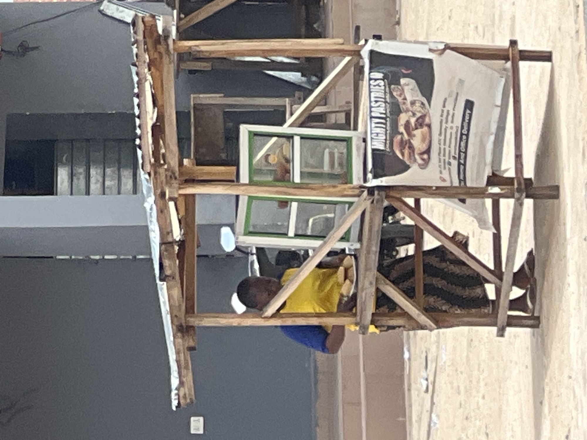 A street vendor is serving pastries from a makeshift wooden stall in Jalingo, Nigeria. The vendor, wearing a yellow t-shirt with blue sleeves and a head covering, is holding a plate of pastries. The stall has a corrugated metal roof and a display case with a green frame. A banner advertising "MIGHTY PASTRIES" hangs on the front of the stall. The ground is sandy and there is a building in the background.