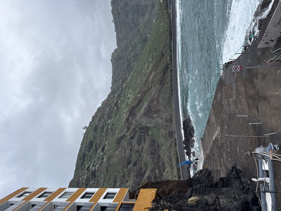 A concrete pier meets a churning ocean beside a steep, green mountain under a cloudy sky. A building with gold and white accents sits in the lower left corner, near black rocks. A road parallels the water, dividing it from the mountain. A "no entry" sign is visible on the pier, along with metal posts and a railing. The perspective is slightly tilted.