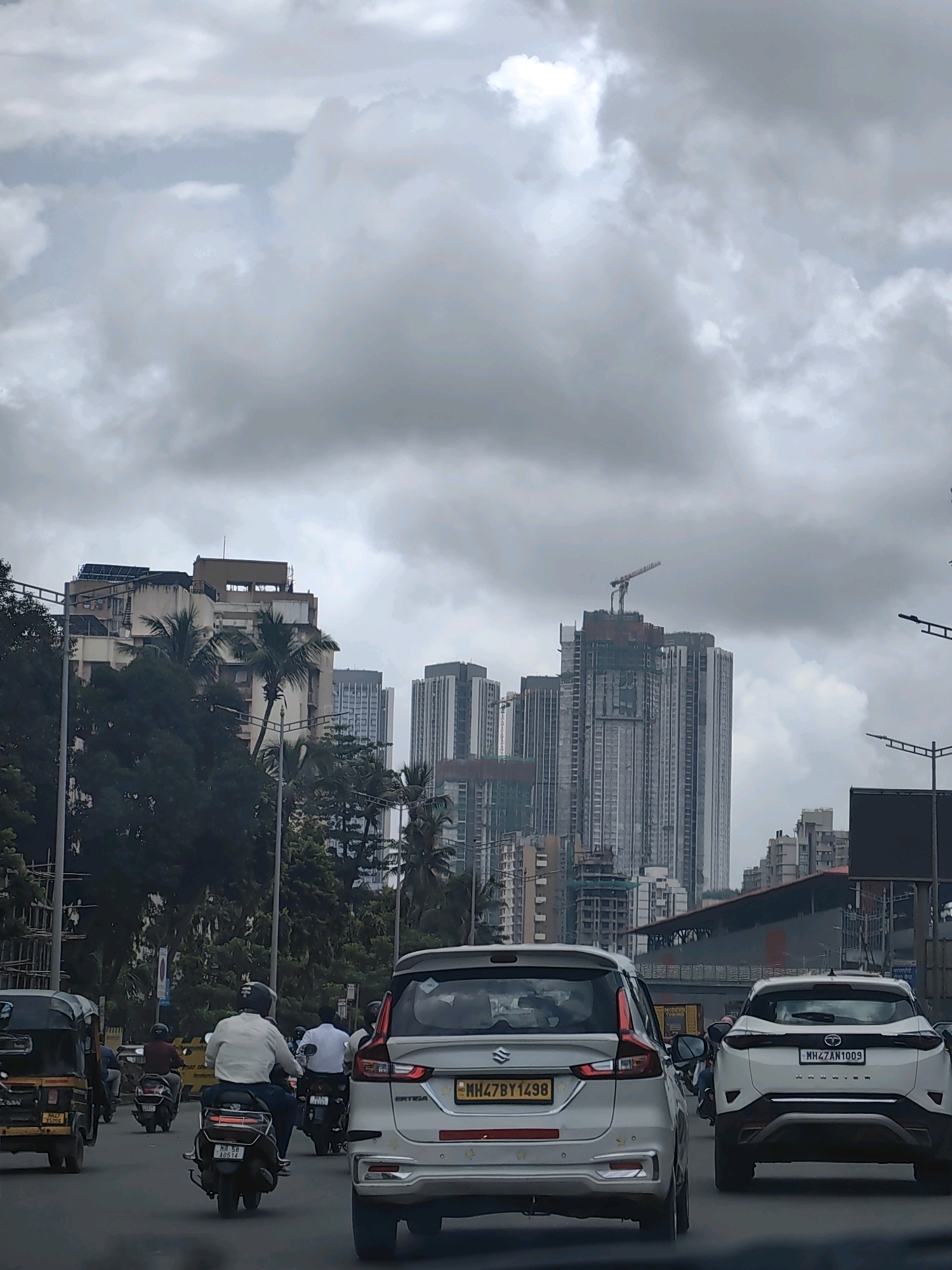 The image depicts a street scene in Mumbai, India, characterized by a cloudy sky over a busy road with vehicular traffic. In the foreground, a silver SUV is prominent, followed by another white SUV. Several motorcycles and a three-wheeled auto-rickshaw are also visible, navigating the road. People are seen riding the motorcycles, some wearing helmets, while others are not. In the background, numerous tall buildings and construction sites dominate the cityscape, some featuring cranes, indicating ongoing development. Green trees and palm trees are interspersed among the buildings, suggesting a blend of urban infrastructure and natural elements. The overall atmosphere is one of a typical bustling day in a major Indian city.