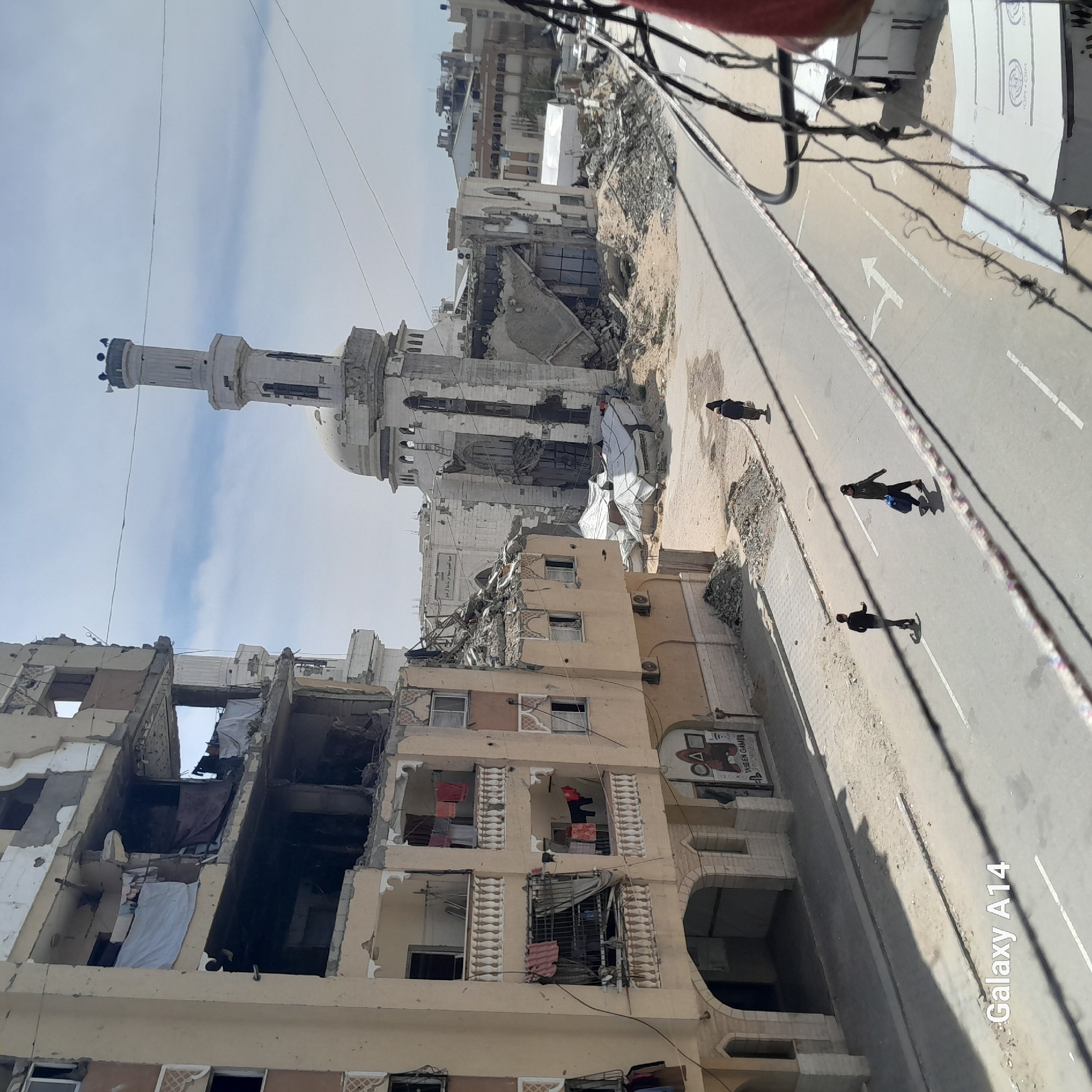 The image depicts a street in an urban environment with significant damage to buildings. A mosque stands prominently with a minaret and dome, also exhibiting damage. Several multistory buildings along the street are partially destroyed, with exposed interiors and hanging debris. Three people are walking on the right side of the road. The road is marked with a directional arrow. A sign is visible on one of the buildings. Overhead wires are strung across the scene.