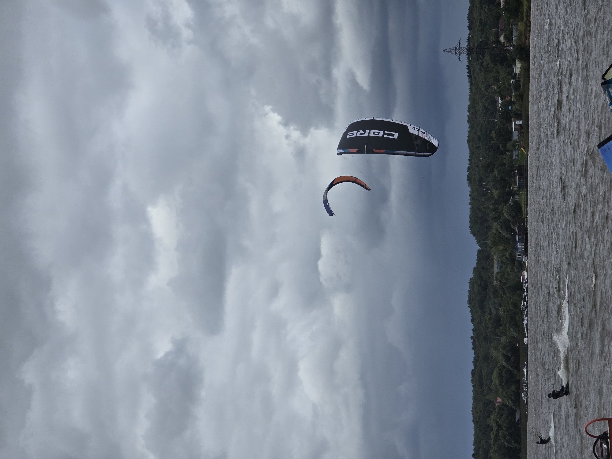 This is an eye-level, medium shot captured on an overcast day in Novoseltsevo, Russia, depicting a scene of watersports on a wide, choppy body of water. The sky is heavily laden with dark, dramatic clouds, suggesting potential rain.
In the middle ground, two individuals are engaged in kitesurfing. One kitesurfer, wearing dark clothing, is positioned to the left, being pulled across the water, leaving a trail of spray. Further to the right and slightly ahead, another kitesurfer is actively riding, also creating a wake. Above them, two kites are visible against the stormy sky: a smaller, orange and blue kite to the left, and a larger black and white kite with the "CORE" logo prominently displayed on the right.
The background features a densely wooded shoreline stretching horizontally across the frame. Along the coast, a scattering of small buildings and structures, possibly houses and boat docks, can be observed. Several boats are moored near the shore. A prominent electricity pylon stands tall on the right side of the background, breaking the uniform line of trees.
In the foreground, partially visible from the bottom edge of the frame, are the elements of what appear to be two more kitesurfing boards or equipment, one blue and black, the other primarily blue. The water surface is rough, characterized by choppy waves and whitecaps. The overall atmosphere is somber and dynamic, dominated by the natural elements of the stormy sky and the active engagement of the watersports participants.