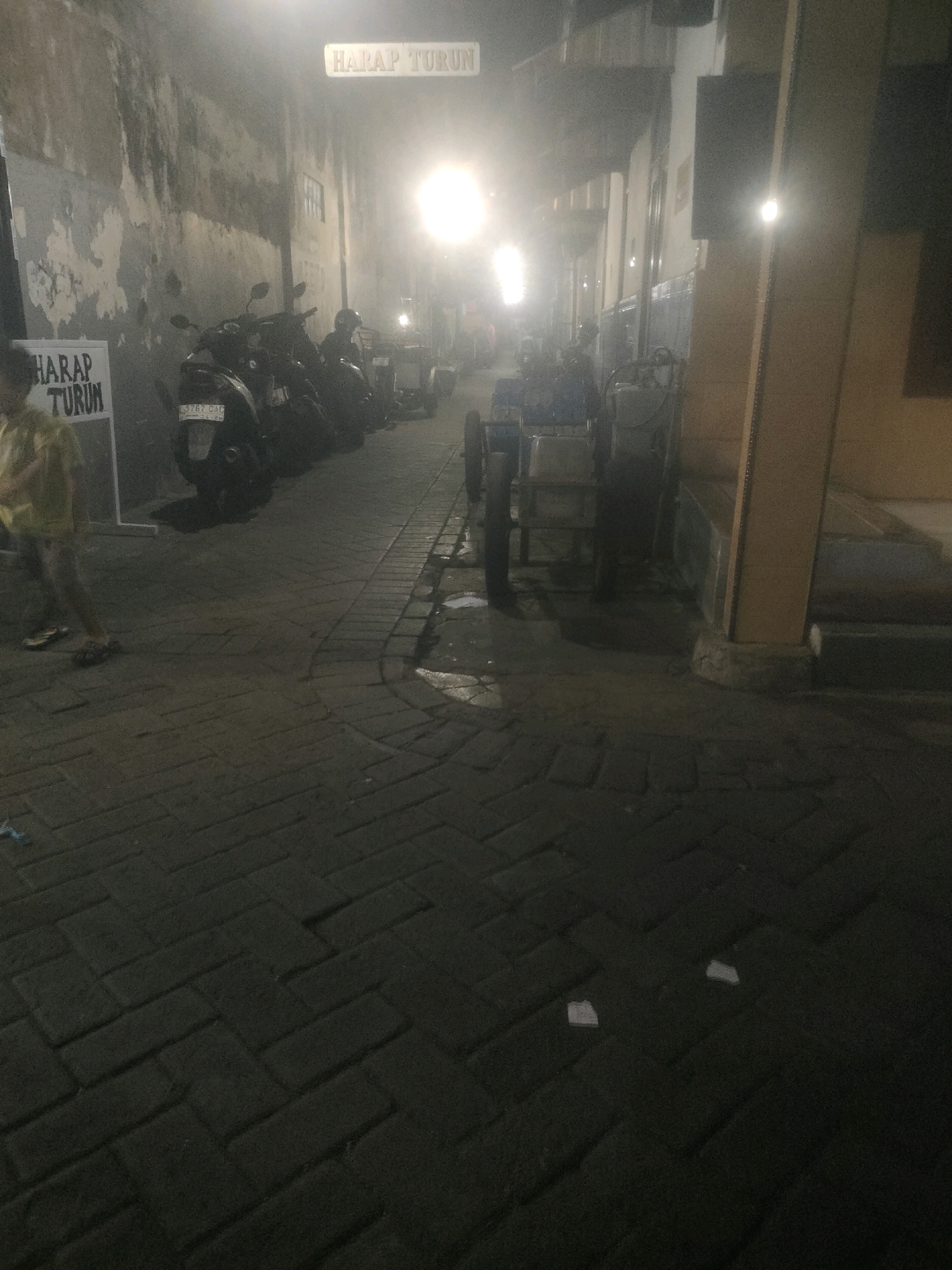 A narrow, cobblestone alleyway in Surabaya, Indonesia, is illuminated by bright lights, creating a hazy atmosphere. Parked motorcycles line the left side of the alley, with a sign that reads "HARAP TURUN" (Please Get Down) hanging above them. On the right, a person in a yellow shirt stands near a white sign with the same inscription. Further down the alley, a cart with wheels is visible. The ground is wet, suggesting recent rain or street cleaning.