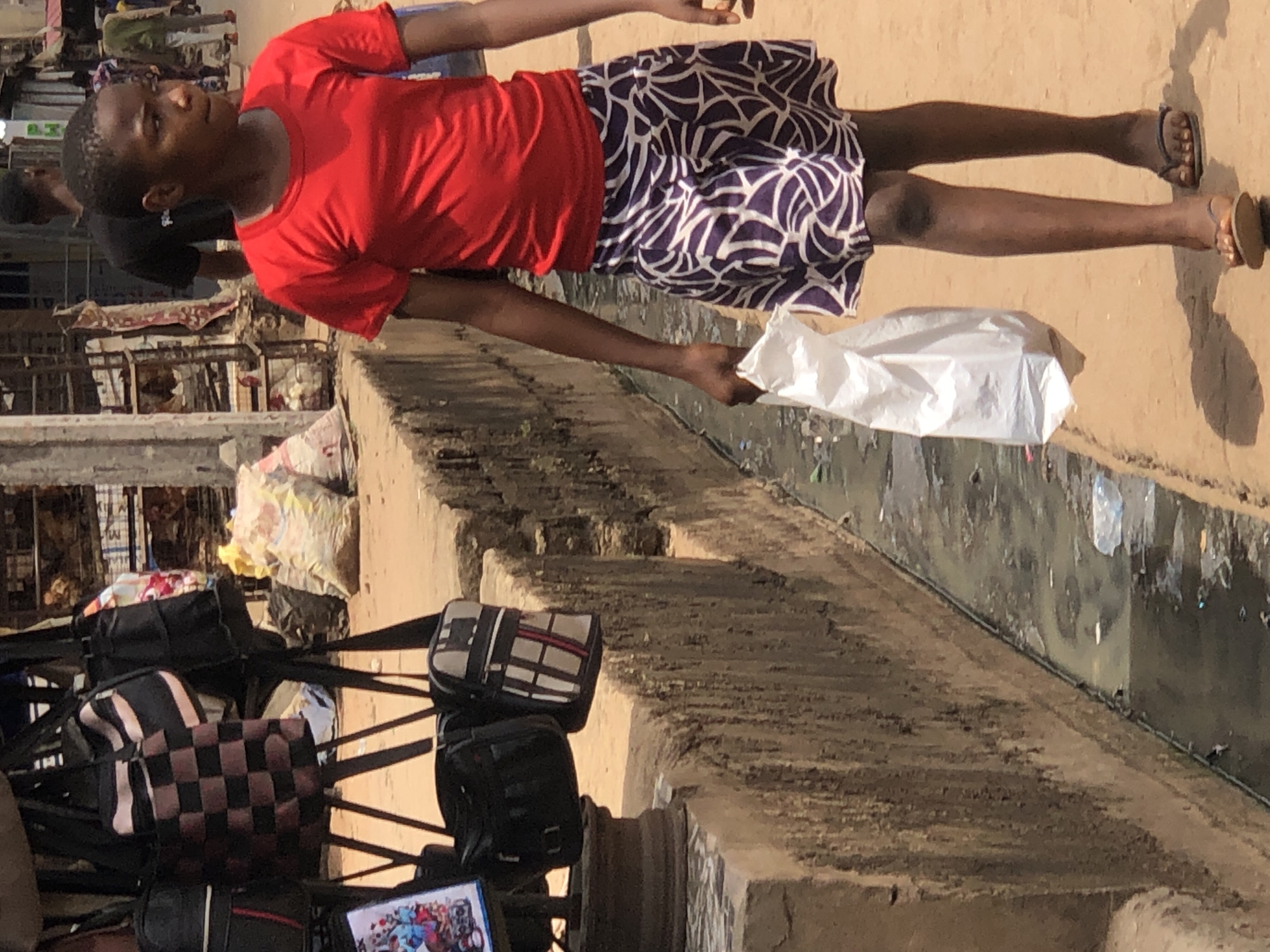 A young person in a red shirt and patterned skirt walks along a street in Onitsha, Nigeria, holding a white plastic bag. The street is lined with open sewers, and bags are displayed for sale.