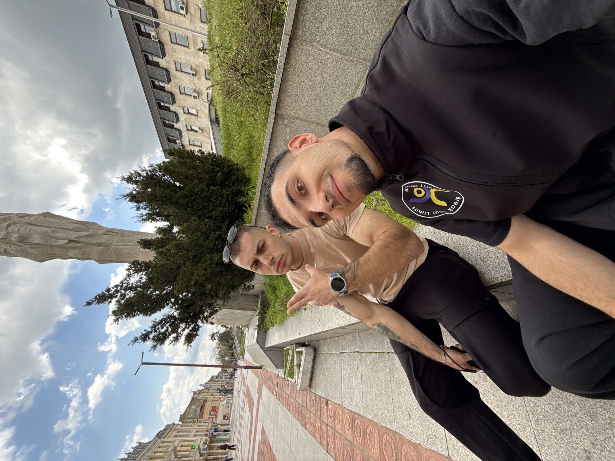 Two young men are posing for a selfie in front of the Monument to 1300 Years of Bulgaria in Shumen, Bulgaria. The monument is partially visible on the left, with a tree in front of it. The men are sitting on a ledge with grass and a low wall behind them. The building behind the wall has multiple windows and air conditioning units. The city street with buildings is visible in the background. The sky is cloudy with patches of blue.