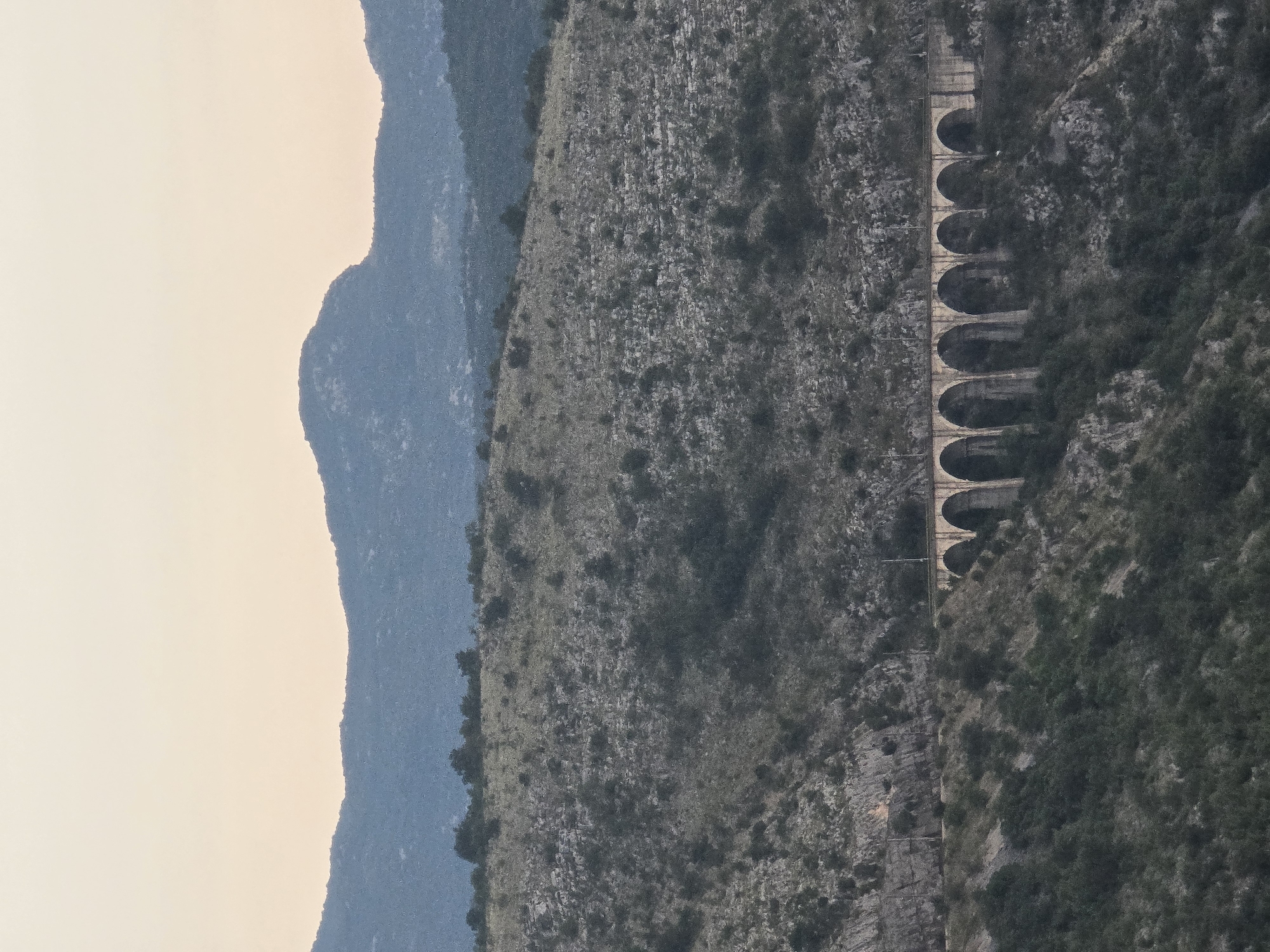 The image shows a stone arched bridge spanning a valley in a mountainous, sparsely vegetated area. The bridge has multiple arches and appears to be part of a railway line, as indicated by the tracks visible on top. The surrounding landscape consists of rocky hills covered in scrub and a few scattered trees. In the background, larger, darker mountains rise against a pale sky. The scene is captured from a distance, giving a sense of scale and isolation. The time of day appears to be late afternoon or early evening, with soft, diffused light and a warm-toned sky. There are no people or animals visible in the image. The location is identified as Klopot, Montenegro.