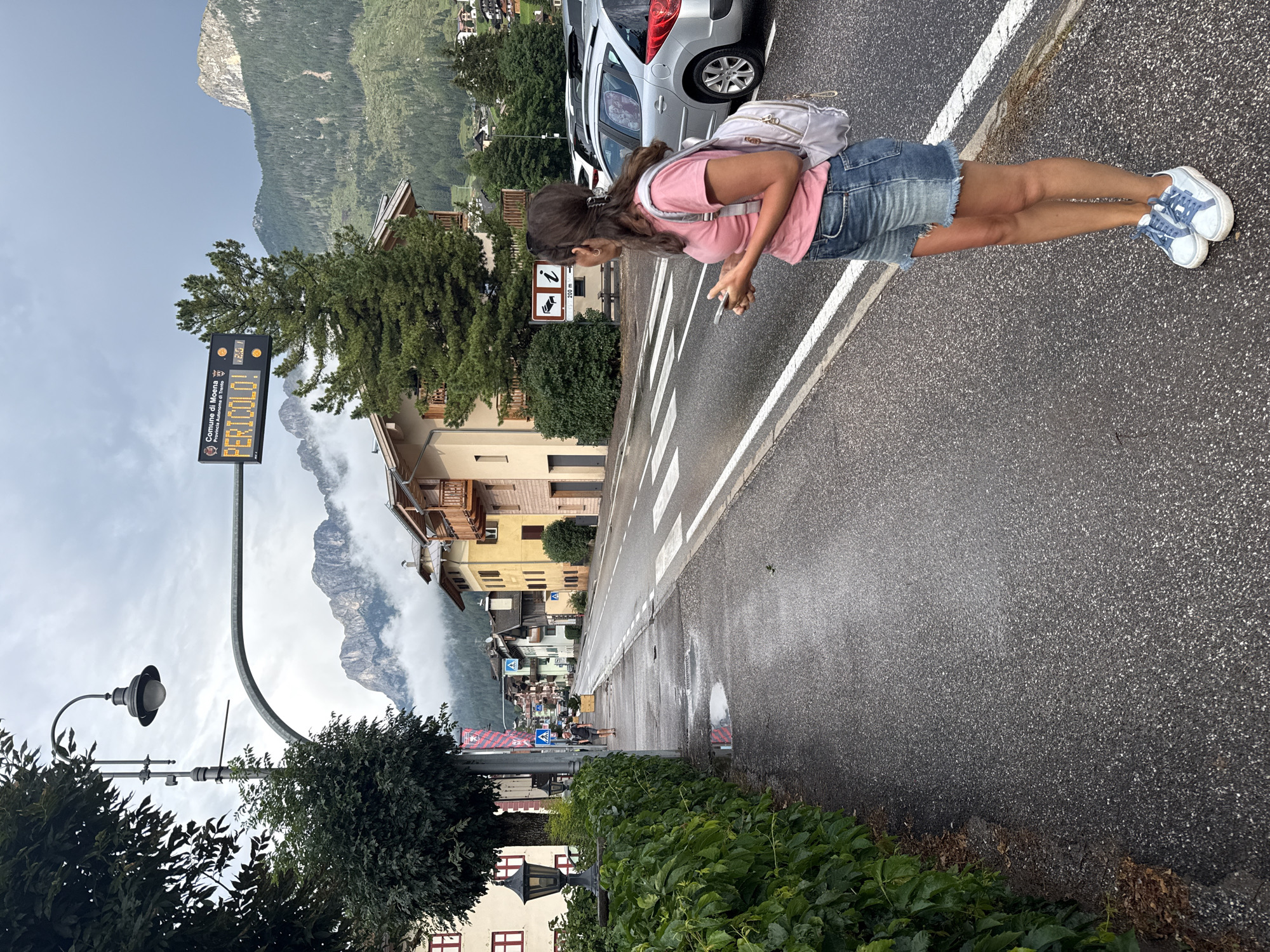 The image shows a street scene in Moena, Italy, under an overcast sky. A young woman with brown hair, wearing a pink t-shirt, denim shorts, and blue and white sneakers, stands on the right side of the street, looking to her right while holding a smartphone. She has a light gray backpack slung over her left shoulder. She is standing on a wet asphalt sidewalk next to a crosswalk. A silver car is parked to her right on the street. Across the street, there are several buildings with colorful facades, including yellow and beige structures with wooden balconies. A large tree with dense green foliage is visible on the left side of the street, bordering the sidewalk. Further down the street, mountains are visible in the background, partially obscured by clouds. A digital sign displaying "COMUNE DI MOENA" and "PERICOLO!" (Danger!) is suspended over the street. A street lamp is also visible on the left side of the image. The street is wet, suggesting recent rain.