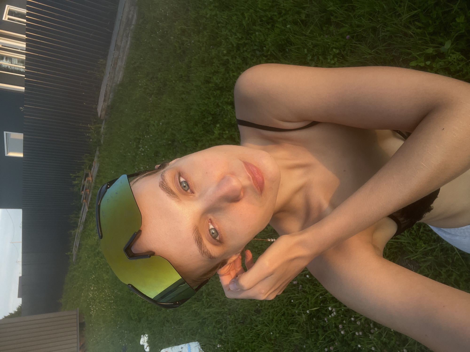The image shows a young woman with blue eyes, wearing oversized green-tinted sports sunglasses. She is outdoors in a grassy area, possibly a backyard, with a dark fence and a building visible in the background. The sun appears to be setting, casting a warm glow on her skin. She is wearing a black bra and gray shorts. Her right hand is raised to her ear, and she is wearing a delicate, dangling earring. The overall scene suggests a casual and relaxed setting in what could be a rural or suburban area outside of a city like Podshivalovo, Russia.