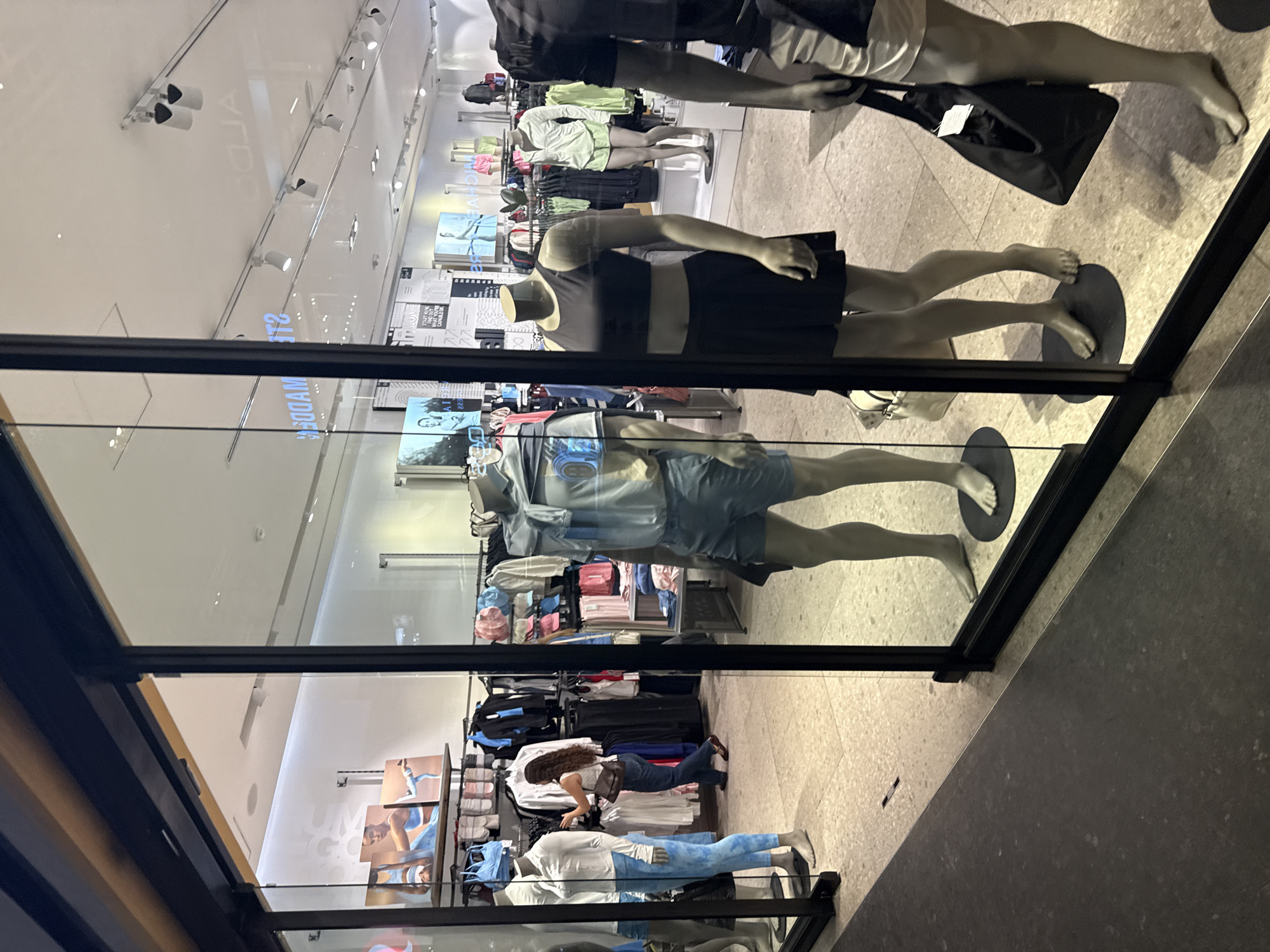 This image captures the interior of a retail clothing store, viewed through a reflective glass display at a tilted angle. The scene is dominated by several mannequins showcasing activewear and casual clothing. In the immediate foreground, a mannequin is dressed in a black tank top and black shorts, while another wears a light blue short-sleeved top and matching shorts. Further inside the store, additional mannequins display a light green activewear top and a white shirt with blue jeans.
Behind the mannequins, a person with long, brown hair, wearing a white shirt, blue jeans, and a dark cross-body bag, is visible. They appear to be interacting with clothing racks, possibly shopping or arranging merchandise. The store's interior features numerous clothing racks laden with various garments in different colors, as well as shelves of folded apparel and wall displays featuring images of models.
The setting is well-lit by artificial ceiling lights, including track lighting. The flooring is a light-colored, speckled material. Reflections on the glass panel superimpose parts of the ceiling and reverse text. Visible text includes "ALDO" reflected on the ceiling, and fragmented, reversed words like "MODAM", "SPORTS", and "VANESSA VINTAGE CANVAS" on display boards inside the store. The image depicts a modern commercial retail environment, characteristic of a shopping mall or similar establishment in Puebla, Mexico, during its operating hours. No specific time of day or weather is discernible due to the indoor, artificially lit setting.