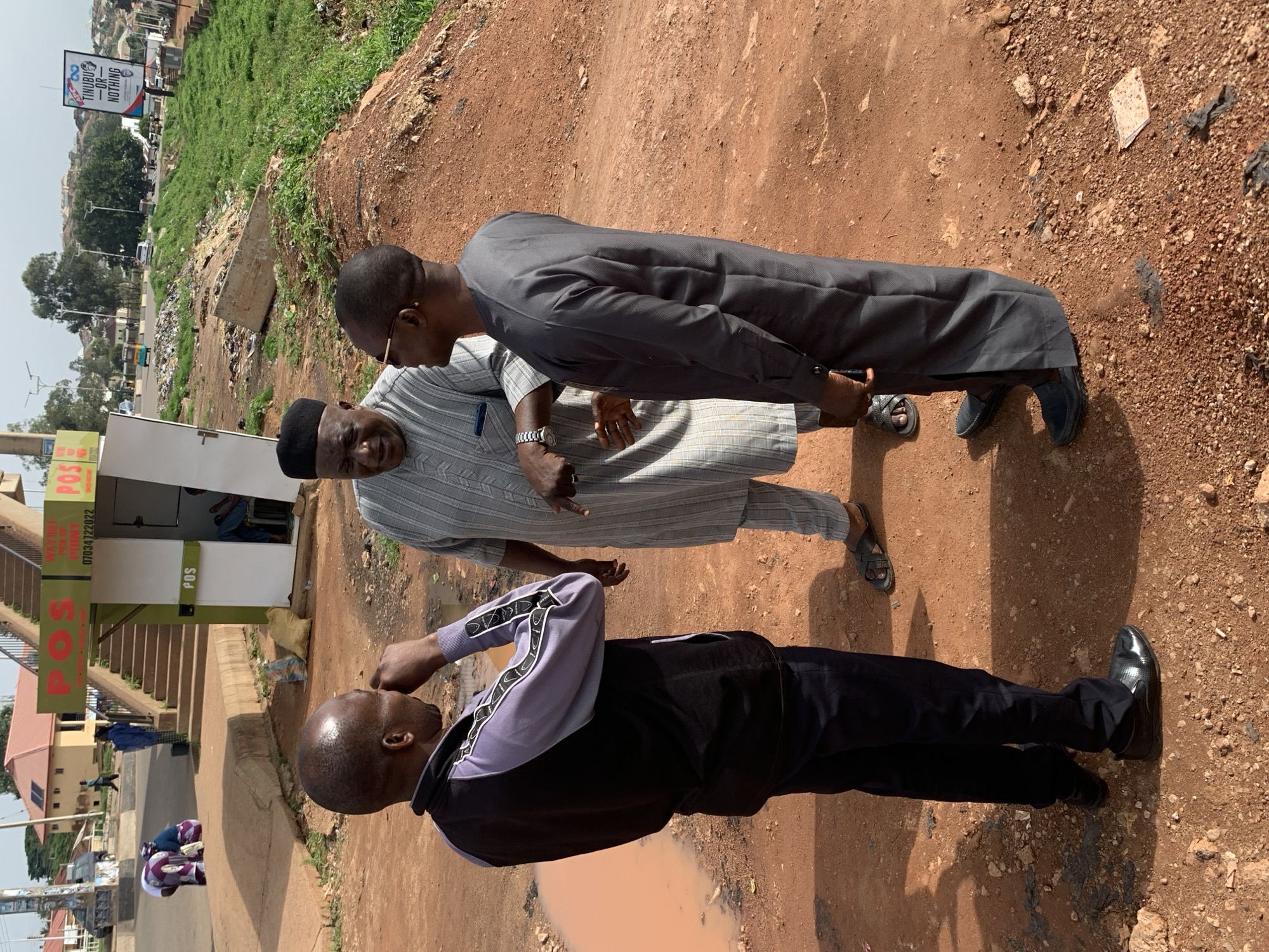 In Gura Top, Nigeria, three men stand in conversation on a dirt path with scattered debris. The man on the left wears a purple and black jacket and dark pants, with his right hand touching his forehead. The man in the center, wearing a striped grey traditional outfit and a black cap, gestures with his left hand while looking at the man on the right. The man on the right, dressed in a grey traditional outfit, stands with his arms down, facing the man in the center. In the background, a "POS" kiosk with stairs leading to a building is visible, along with a billboard that reads "TINUBU - OR - NOTHING." A muddy puddle is present on the left side of the path. The environment suggests an urban area with signs of development and neglect.