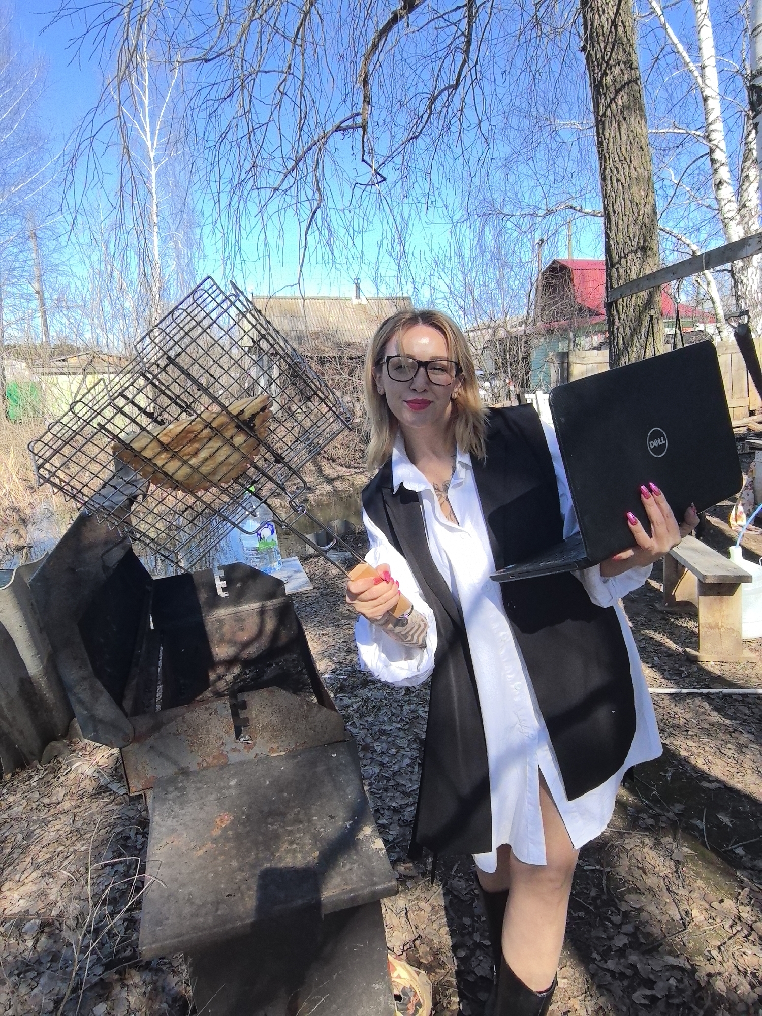The photograph depicts a woman in an outdoor setting, likely a backyard or rural area. The woman, who has blonde hair and is wearing glasses, is holding a Dell laptop in her right hand and a grilling tool in her left. She is dressed in a white shirt and a black vest, with black boots completing her attire. A grill is positioned in front of her, with a metal grate holding what appears to be fish being cooked. The background features a mix of trees, some with bare branches, and buildings, including a red-roofed house. A table, a water bottle, and other unidentified objects are scattered around the scene. The overall setting seems to be a sunny day in a possibly suburban or rural location near Noviy, Russia, given the context provided.