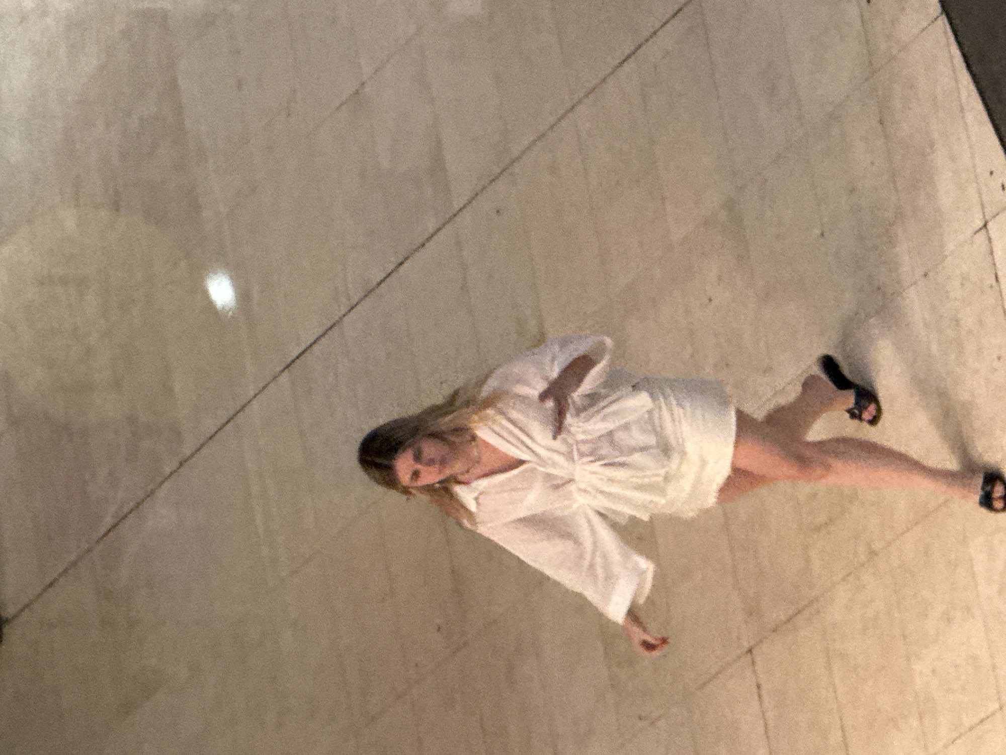A young woman with blonde hair walks across a tiled floor. She is wearing a white collared shirt with loose sleeves, tied at the waist with a belt, and a white mini-skirt. She also wears black sandals. The floor is made of large, light-colored tiles with dark grout lines, and a diagonal line is visible across the tiles. The scene appears to be indoors, possibly in a shopping mall or public building in Barcelona, Spain.