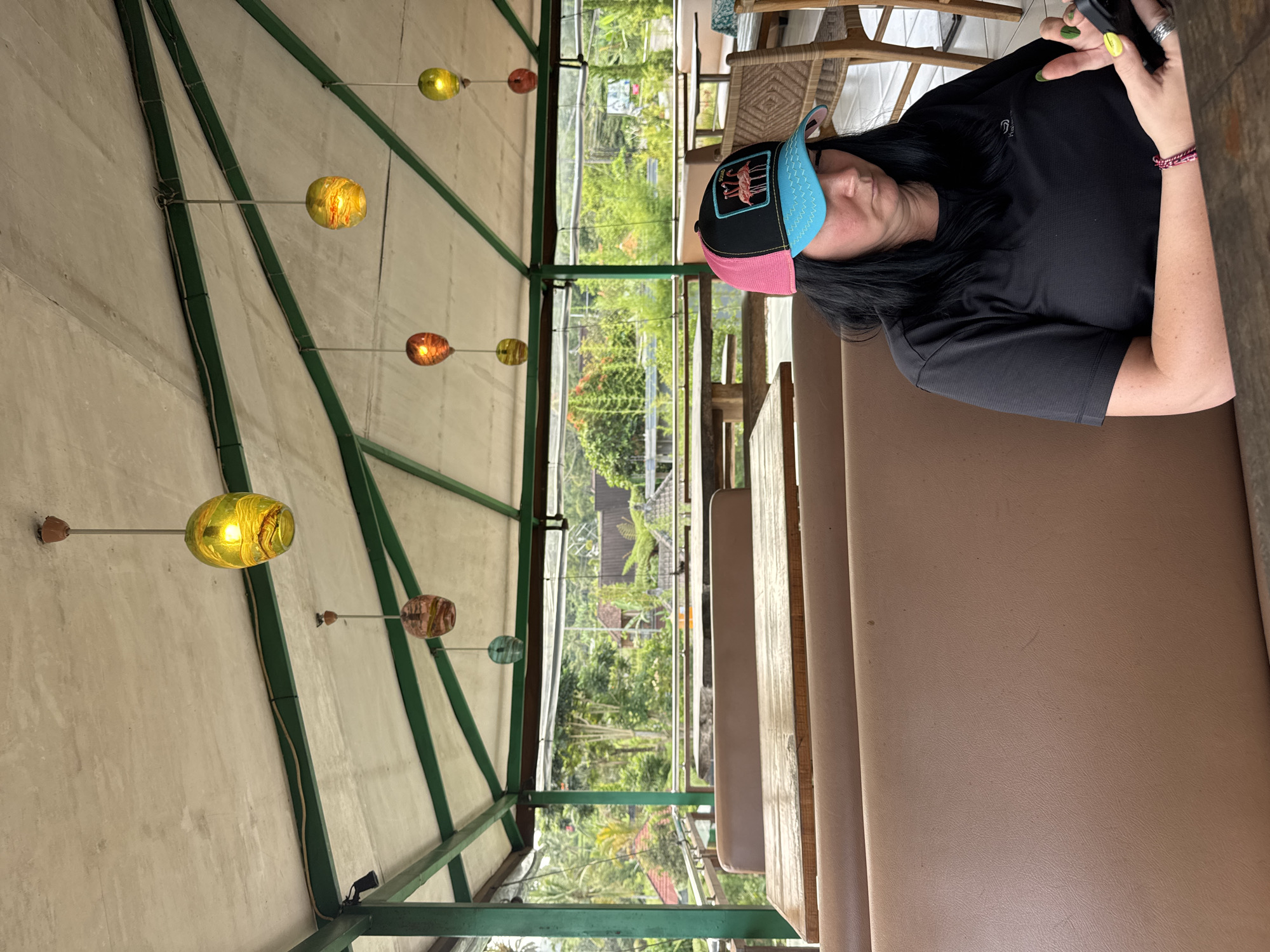 A woman is seated at a table on a bench under a pavilion-like structure in Baturiti, Indonesia. The pavilion has a concrete roof supported by green metal beams. Hanging from the ceiling are glass lamps. The surrounding area is lush with greenery. The woman is wearing a pink and blue cap, a black t-shirt, and she appears to be holding a mobile phone.