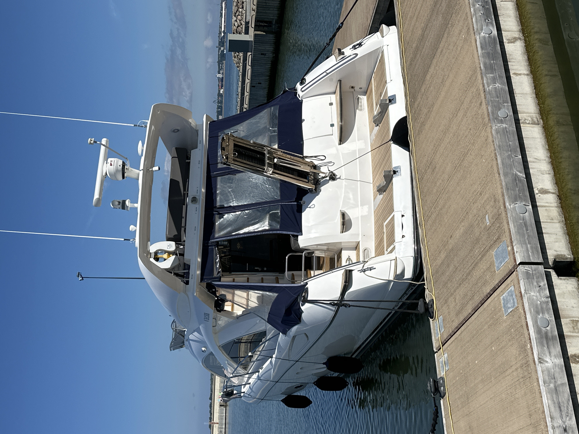 A white luxury yacht, identified by the registration number V26133, is docked at a wooden pier in Tallinn, Estonia. The yacht is white with navy blue canvas covers on its aft section. Its stern features a wooden deck with two sets of chocks. Three black fenders hang from the yacht's starboard side. The yacht is secured to the pier by black ropes to black bollards. The water is calm and appears to be greenish. In the background, a breakwater constructed of rocks and concrete structures is visible, along with industrial cranes and buildings in the distance, suggesting a port or harbor area. The sky is clear and blue with a few wispy clouds. There are no people visible in the image.