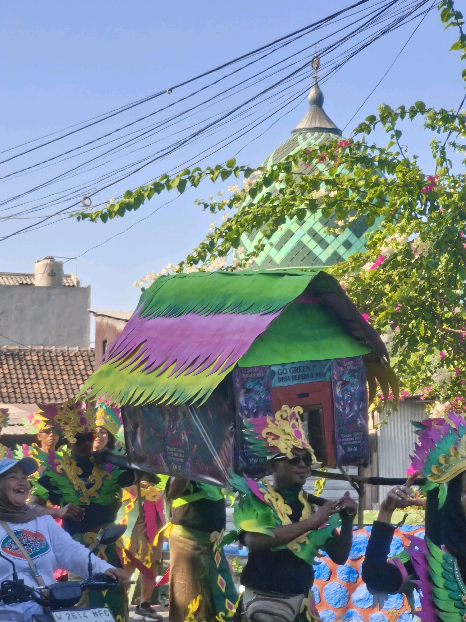 In the city of Kepuh Kiriman, Indonesia, a vibrant parade unfolds under a clear blue sky. The focal point is a large, colorful float resembling a house, adorned with green, pink, and yellow fabric resembling leaves or feathers. The float is carried by several individuals dressed in elaborate, festive costumes featuring golden headdresses and green and yellow attire. One person in the foreground, seated on a motorcycle, is wearing a white long-sleeved shirt and a light brown hijab, smiling towards the camera. Another person in a costume is visible on the right, with their hand raised. In the background, a distinct green domed structure, possibly a mosque or a unique architectural element, is visible above the trees. Overhead, a network of power lines crisscrosses the sky, a common sight in urban and semi-urban Indonesian landscapes. The scene captures a lively community event, likely a religious or cultural celebration, marked by traditional dress and elaborate decorations.