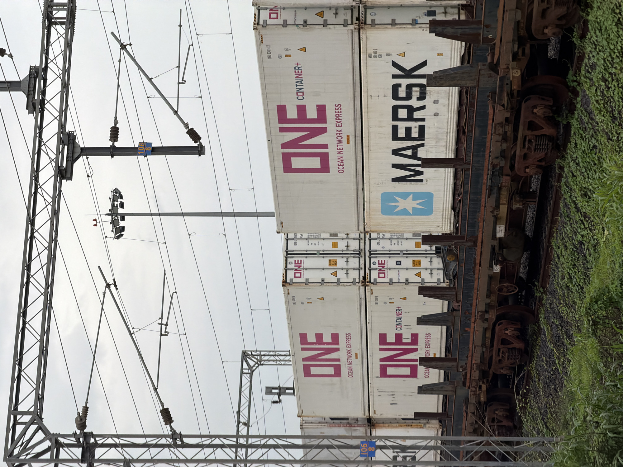 The image captures a freight train loaded with shipping containers, passing through Saphale, India. The train features multiple stacked containers. The top row of containers displays the "ONE OCEAN NETWORK EXPRESS" logo in large pink lettering. Below these, a container with the "MAERSK" logo is visible. The train cars are old and rusty. Overhead, a complex network of electrical wires and support structures for an overhead catenary system is present, indicating the railway is electrified. The sky is overcast. The surrounding area appears to be a railway embankment with sparse green vegetation.