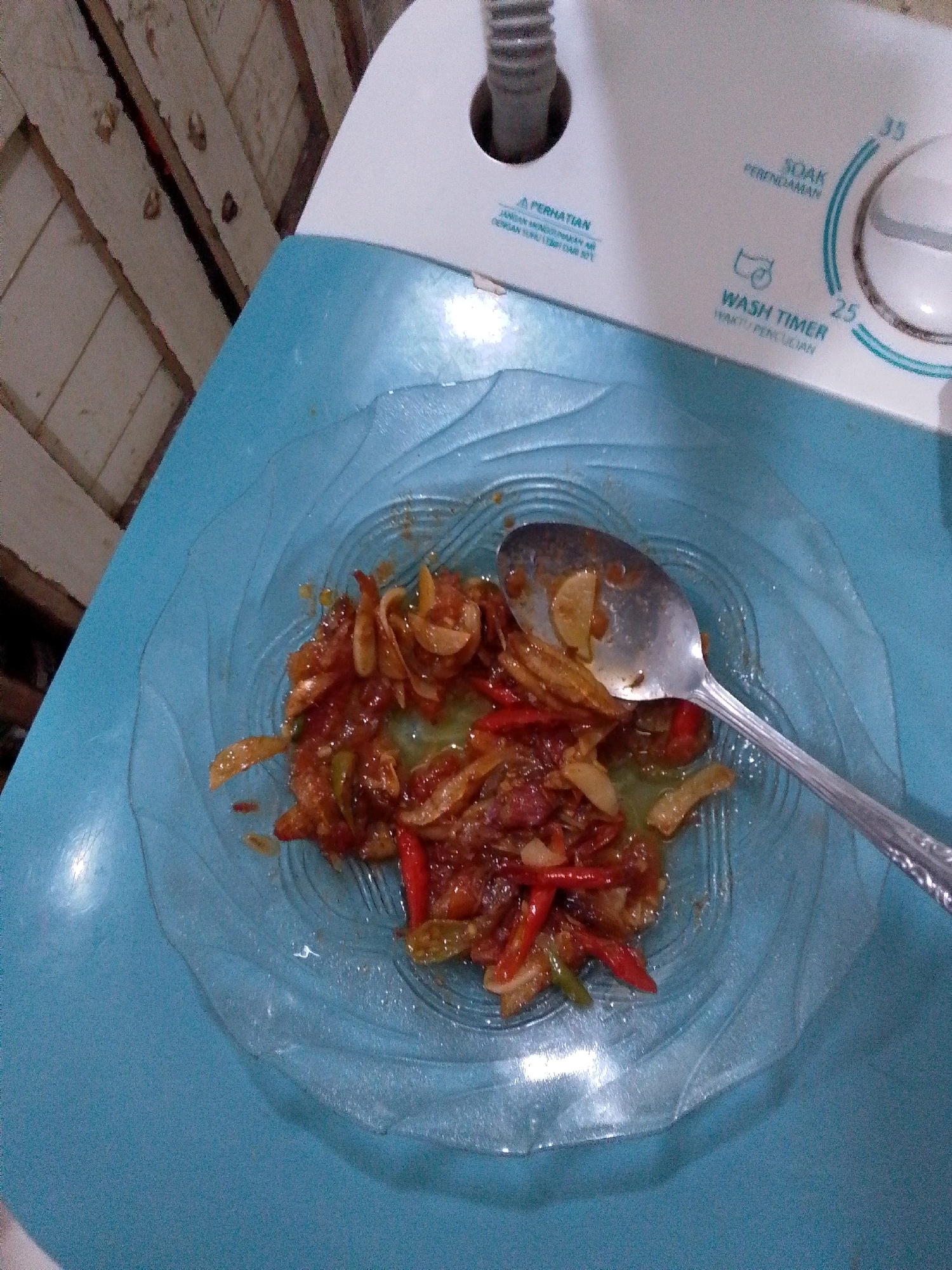 The image shows a close-up of a plate of food on a blue surface, which appears to be the lid of a washing machine. The food is a spicy stir-fry, likely containing chilies, onions, and possibly some form of protein, all coated in a reddish sauce. A metal spoon rests on the plate, partially submerged in the food. The background shows a wooden wall and part of the washing machine's control panel, which has labels in Indonesian, including "SOAK" and "WASH TIMER." The scene is likely captured in a domestic setting, possibly in East Jakarta, Indonesia, given the language on the appliance.